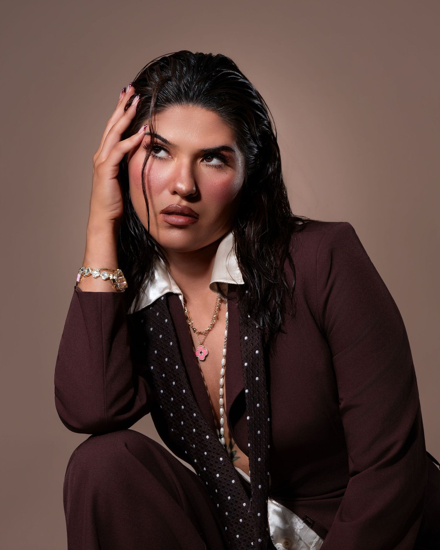 Woman in brown suit with wet hair poses thoughtfully, wearing gold jewelry and a pink necklace against a tan background.