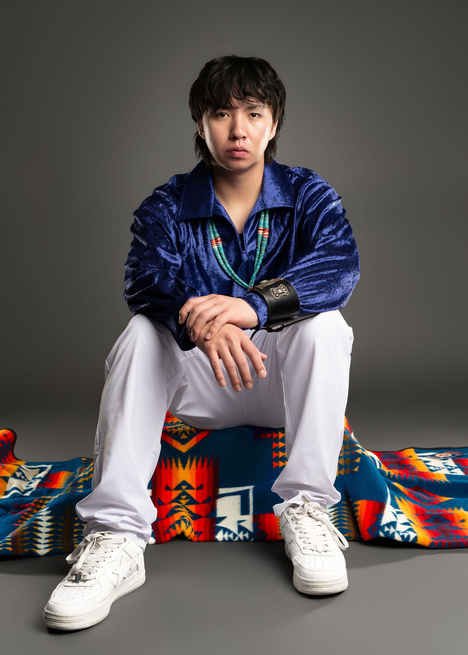 Person in blue velvet jacket and white pants sitting on a colorful Pendleton blanket in a studio.