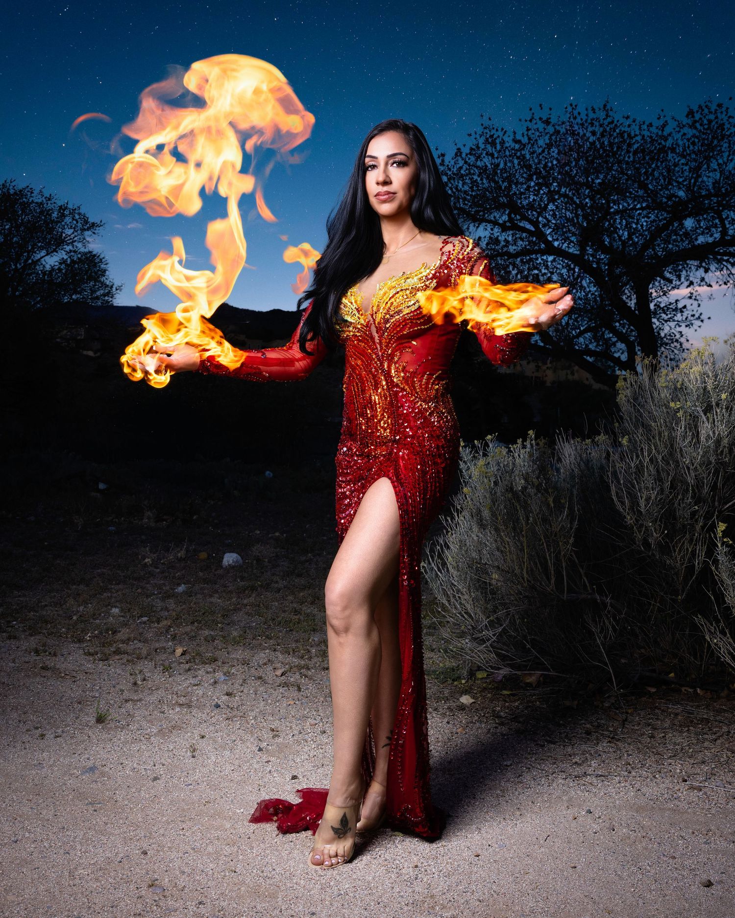 Woman in red sequin gown conjuring flames in desert night setting.