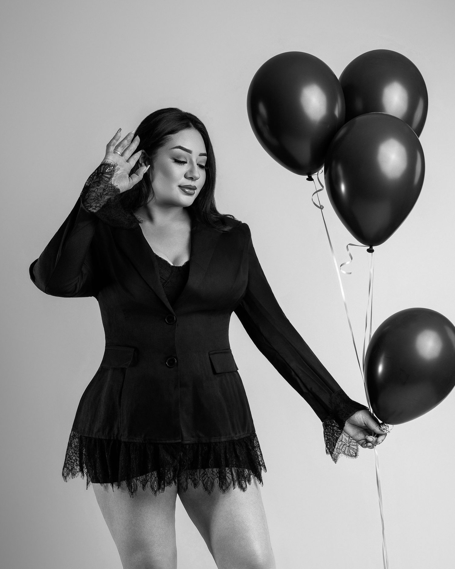 Woman in black blazer and lace shorts holds dark balloons, posing elegantly in a black and white photo.