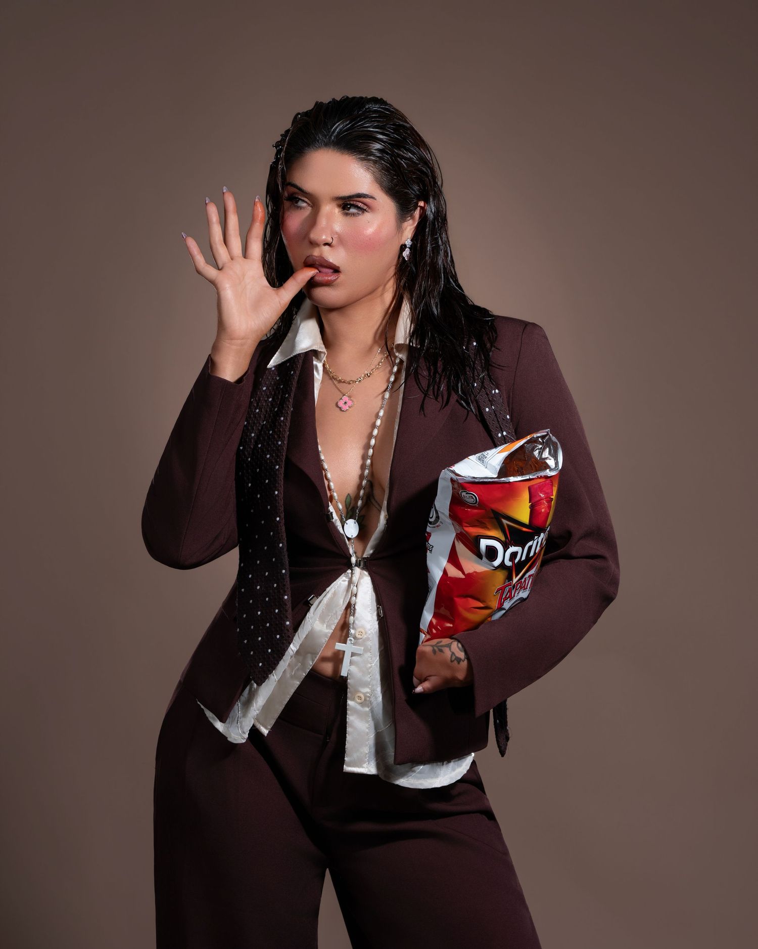 Woman in brown suit holding oversized Doritos bag, licking fingers with surprised expression.