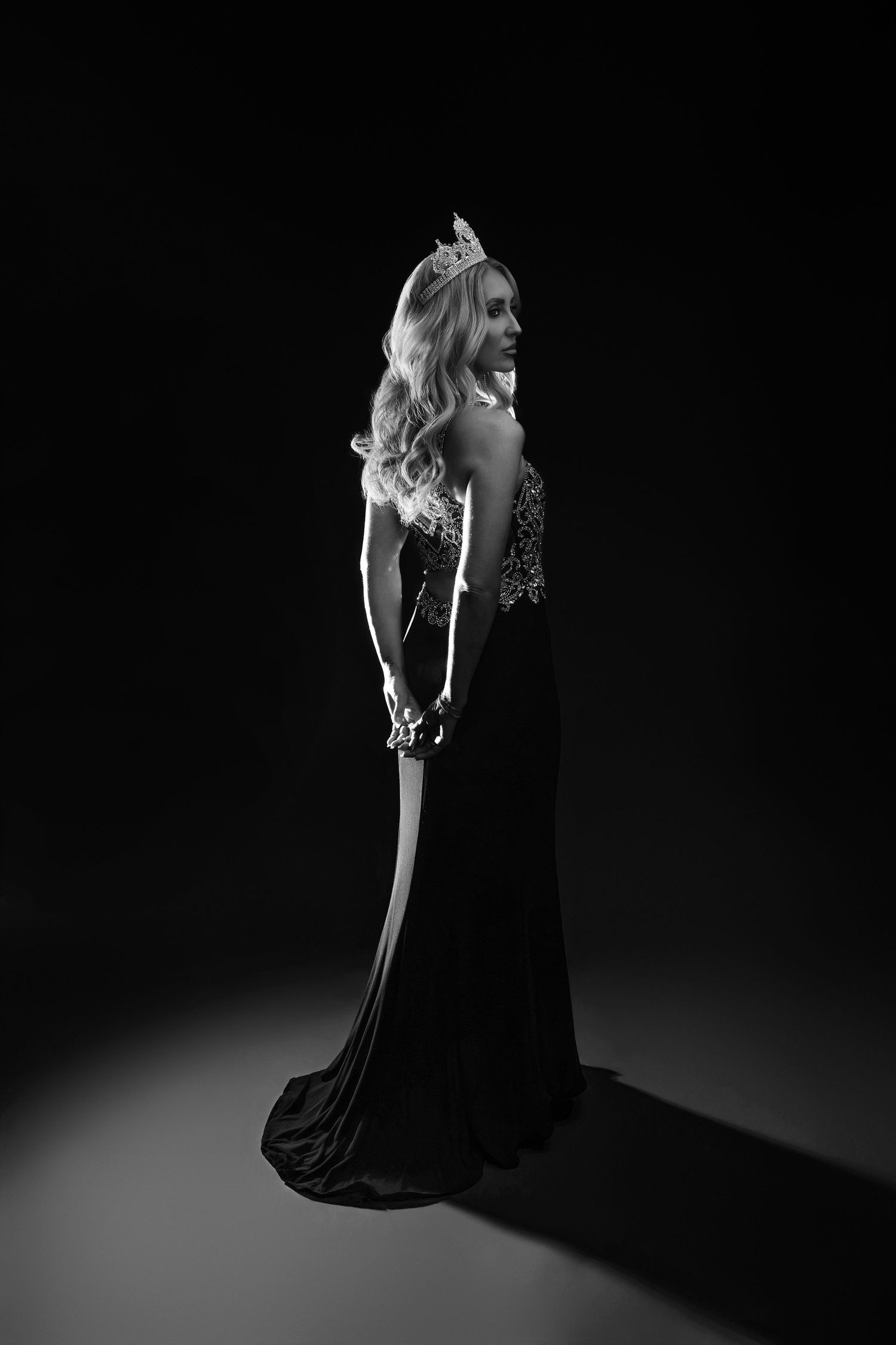 Black and white photo of a woman in an elegant gown and crown posing gracefully in a dark studio setting.