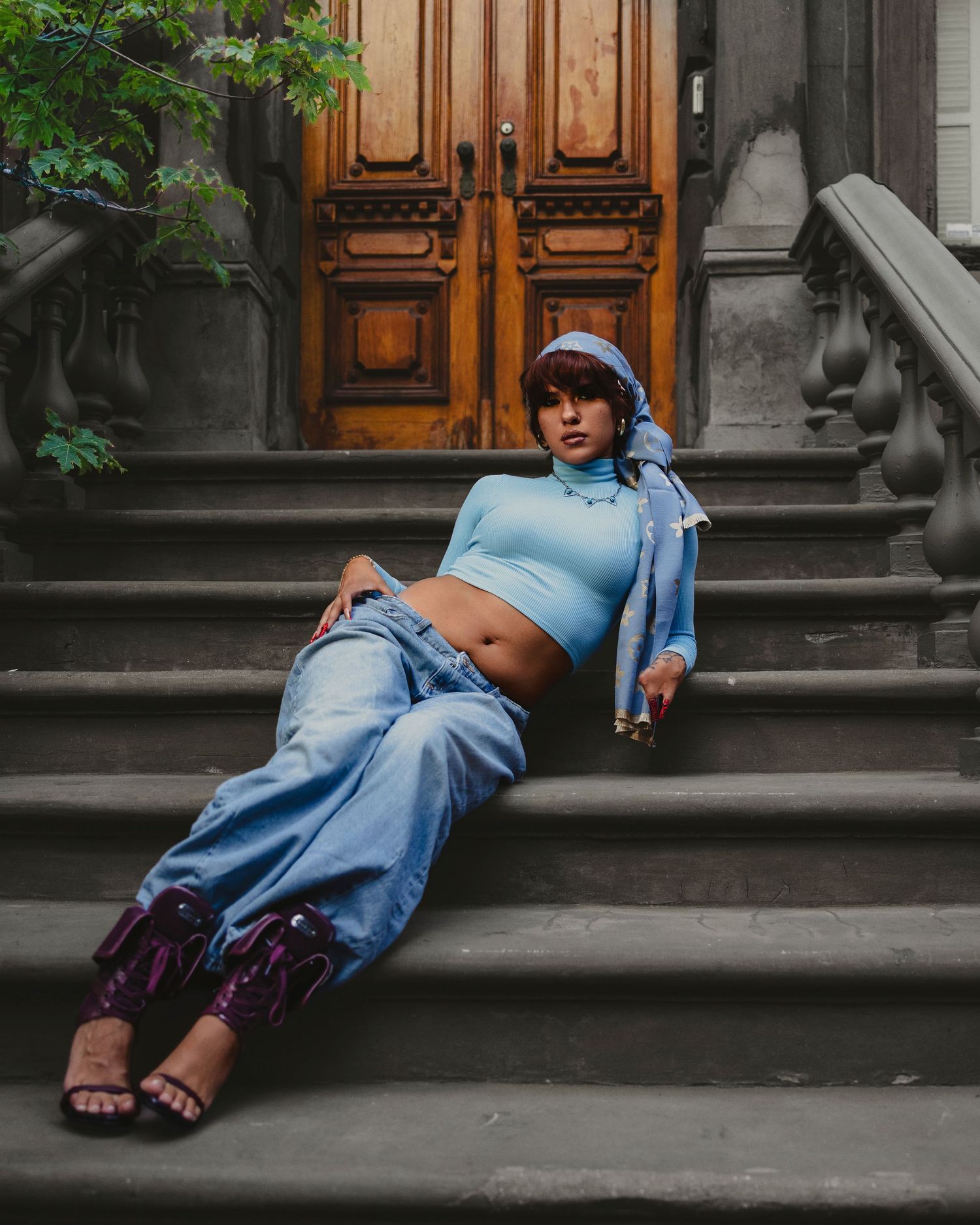 Woman in blue crop top, wide-leg jeans, and purple boots poses on stone steps before a wooden door.