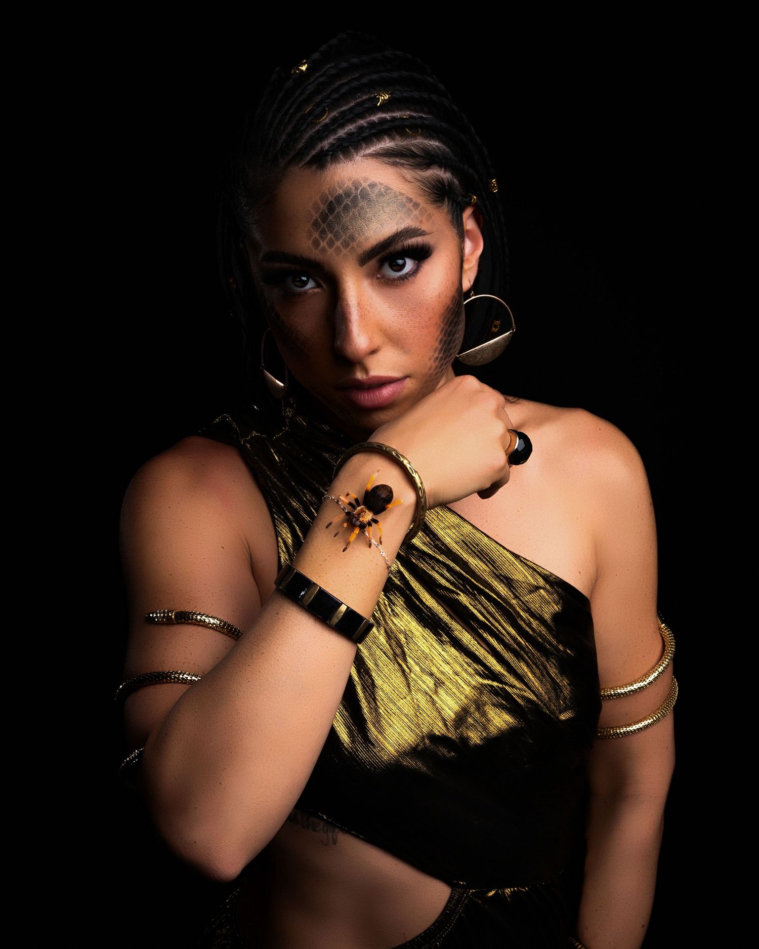 Woman in gold fringe top with braids, hoop earrings, and gold bangles poses against black background.
