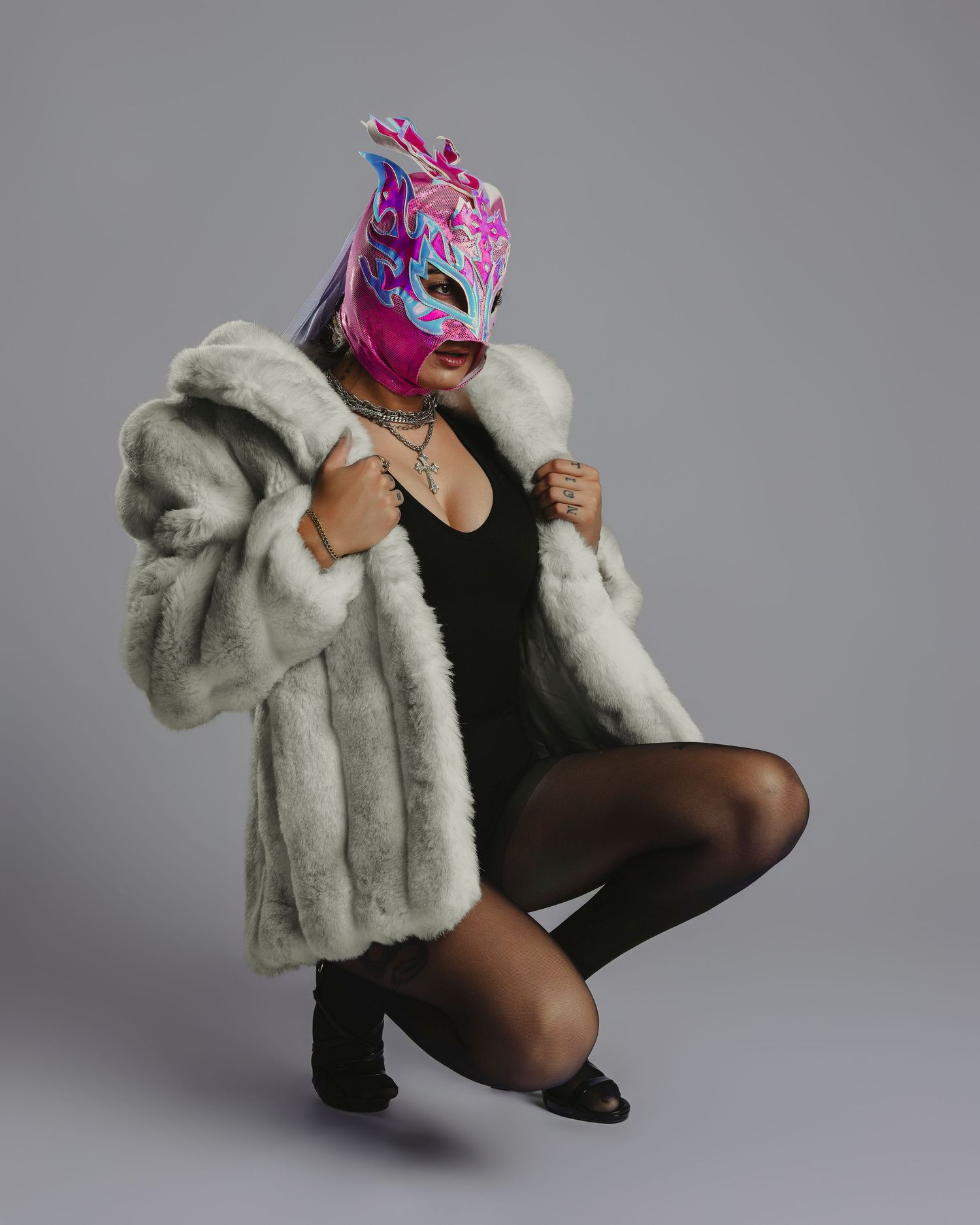 Person wearing colorful lucha libre wrestling mask and white fur coat posing dramatically in black bodysuit and tights.
