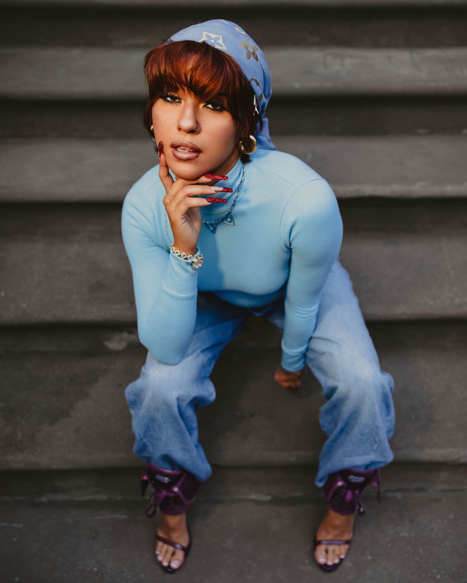 Woman in blue bodysuit, patterned headscarf, and wide-leg jeans poses on gray steps wearing purple heeled sandals.