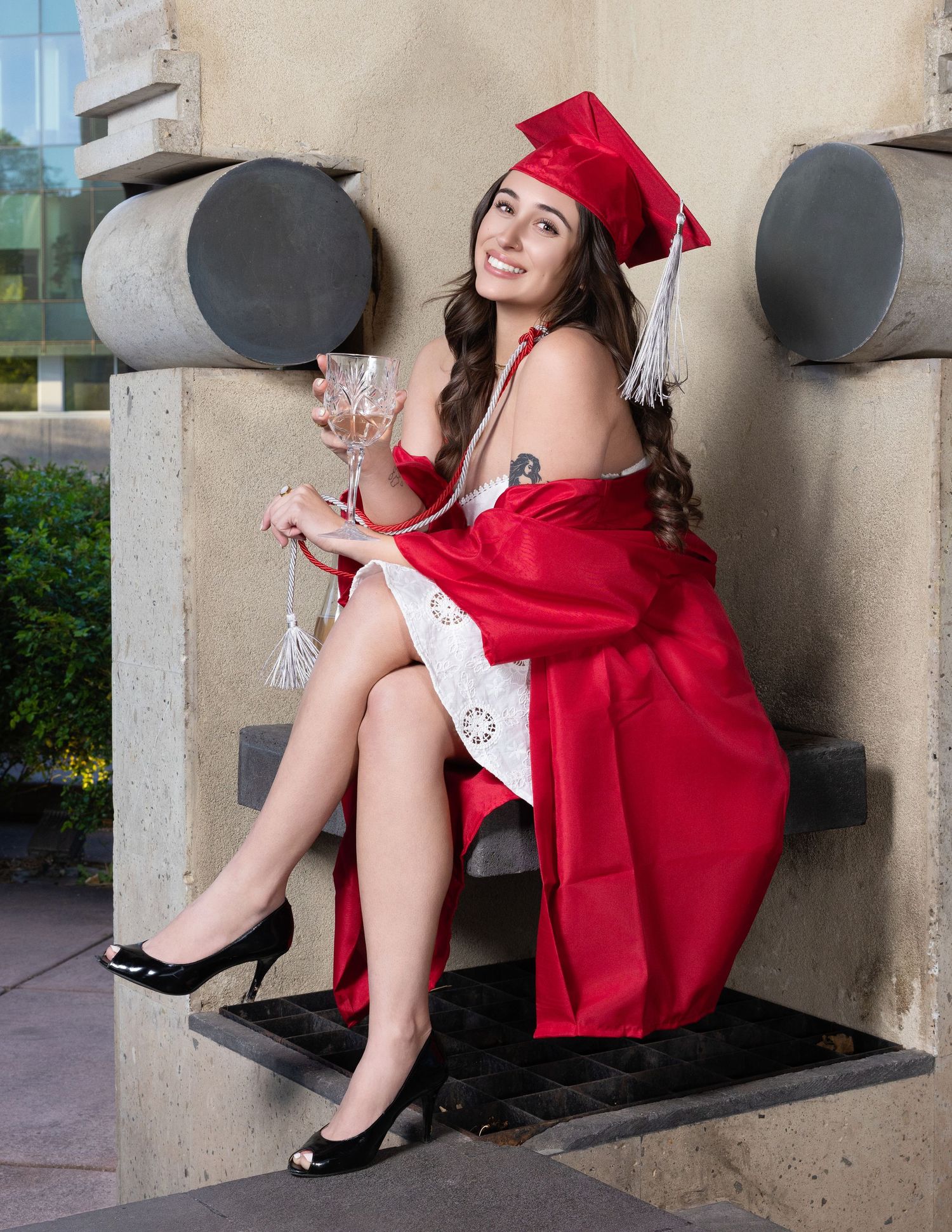 Graduate in red cap and gown holds wine glass while seated on stone steps outdoors.