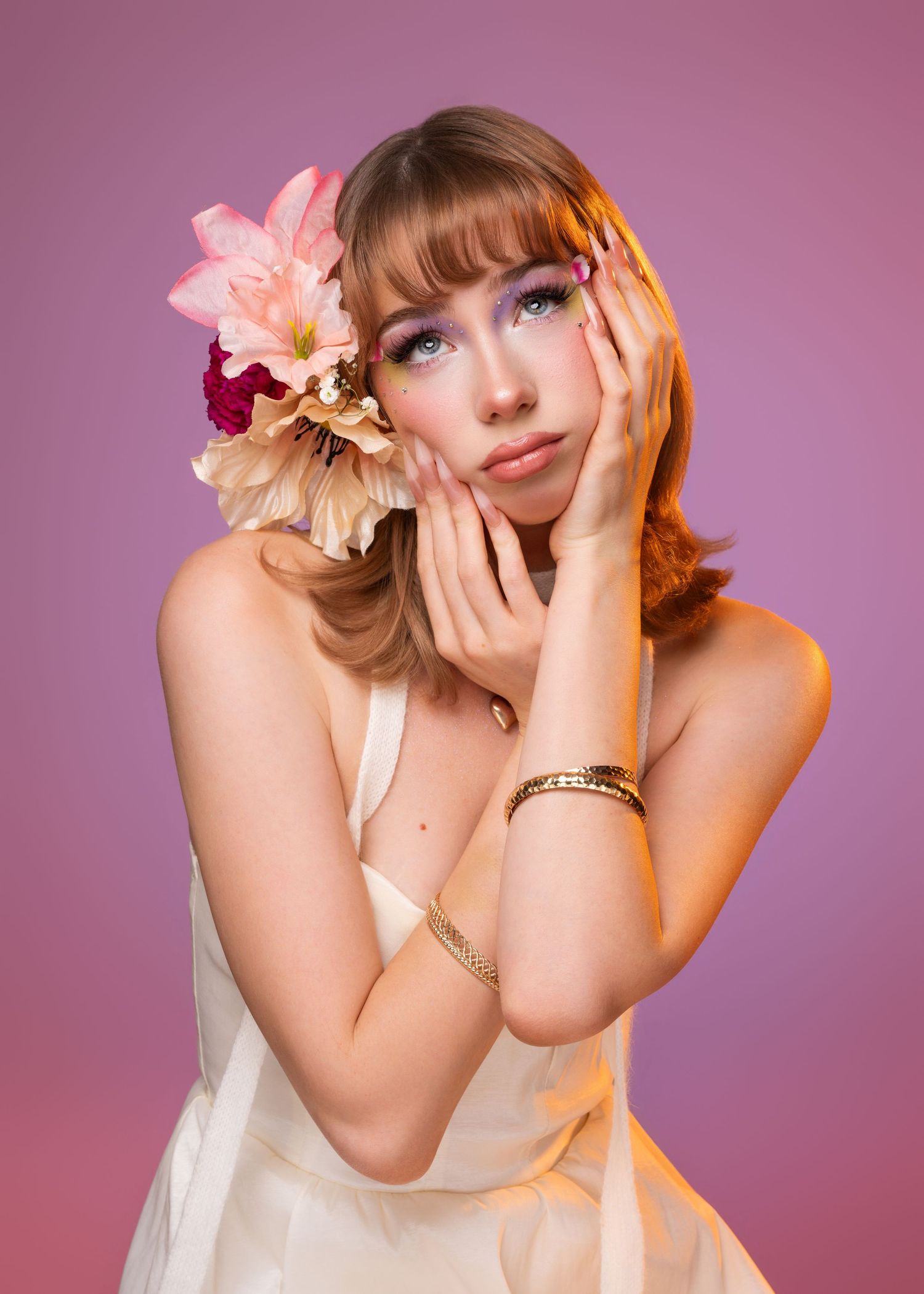Young woman with red hair, pink flowers, gold bracelet, white dress, posing against a pink-purple gradient background.