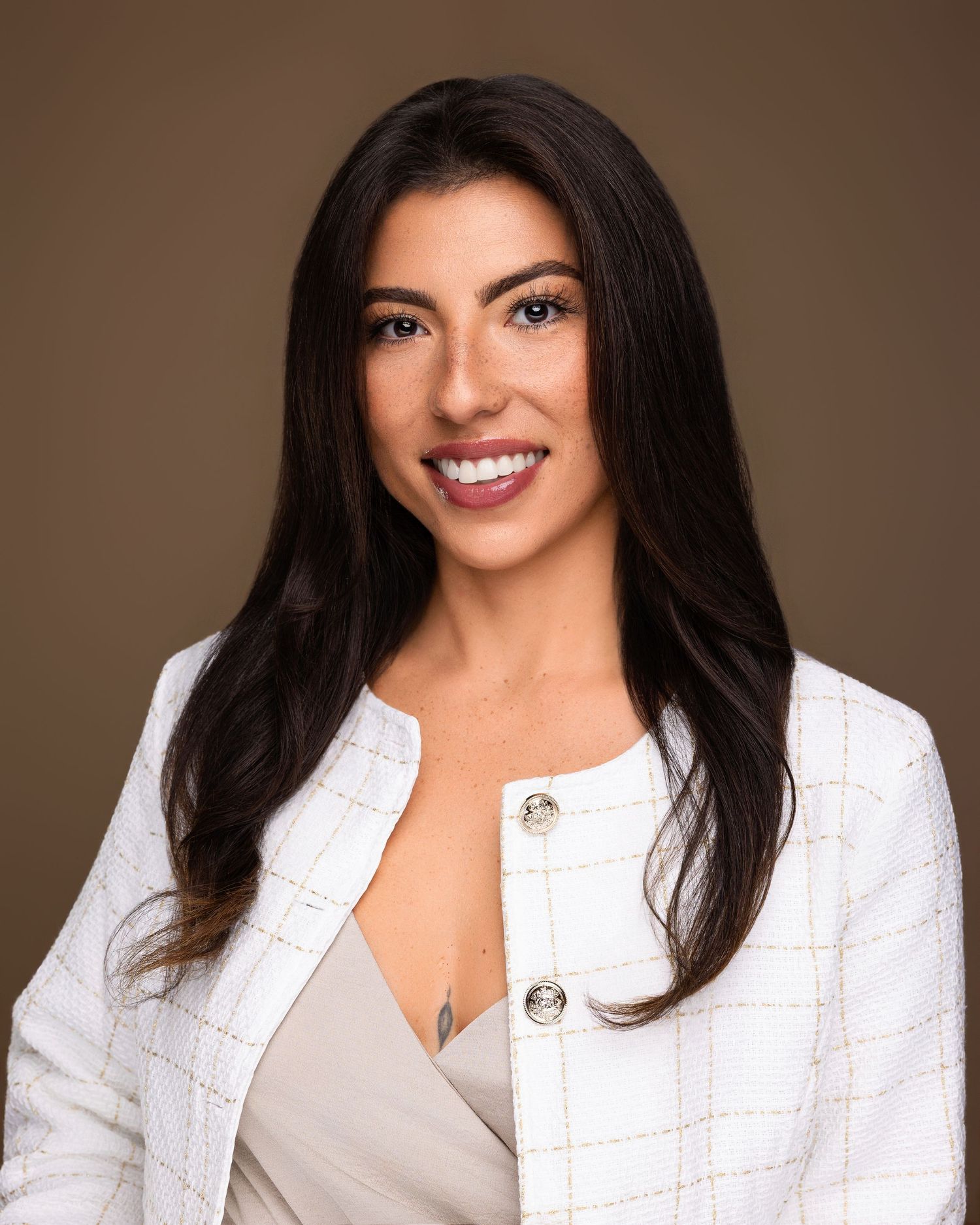Professional headshot of a smiling woman with long dark hair wearing a white blazer against a brown background.