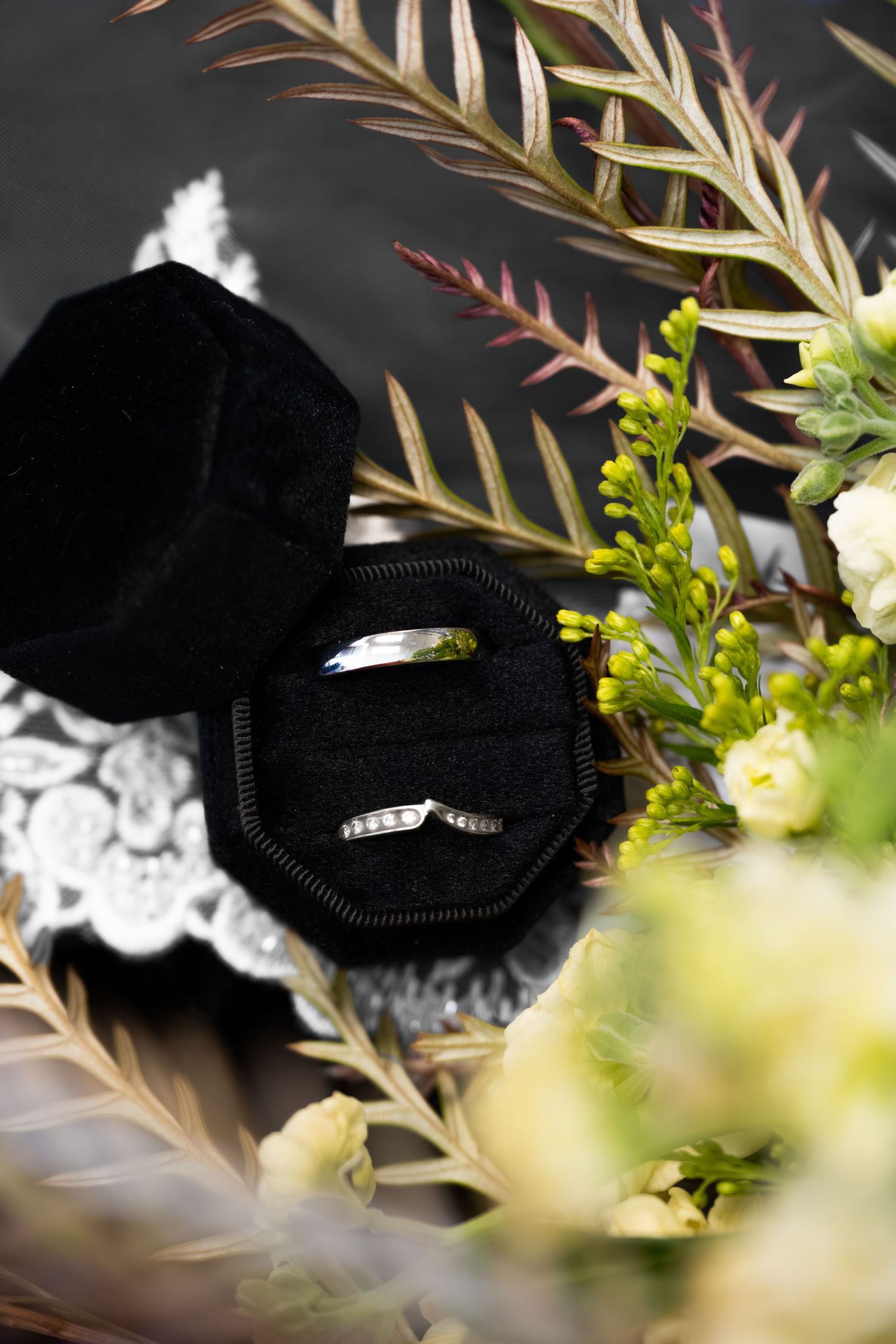 Silver wedding band set in black velvet ring box surrounded by greenery and white floral arrangement.