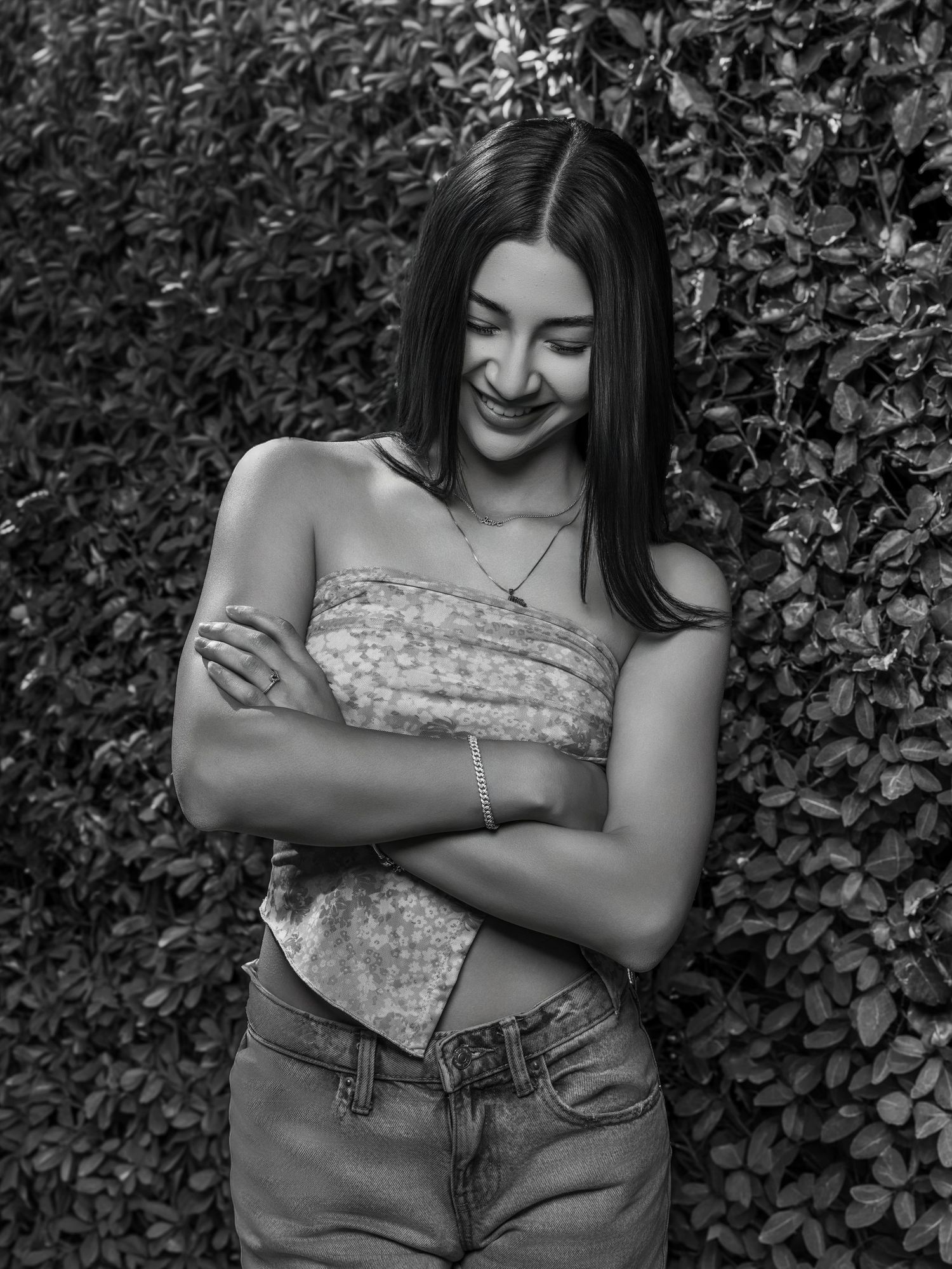 Young woman with arms crossed smiles looking down against green leafy wall in black and white photo.