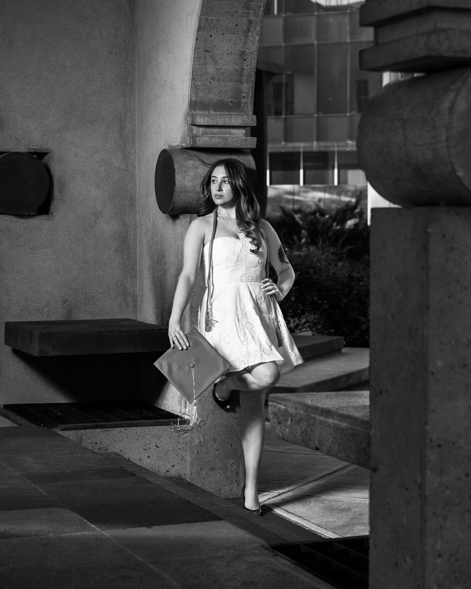 Black and white photo of a young woman in a white dress holding a graduation cap outdoors.