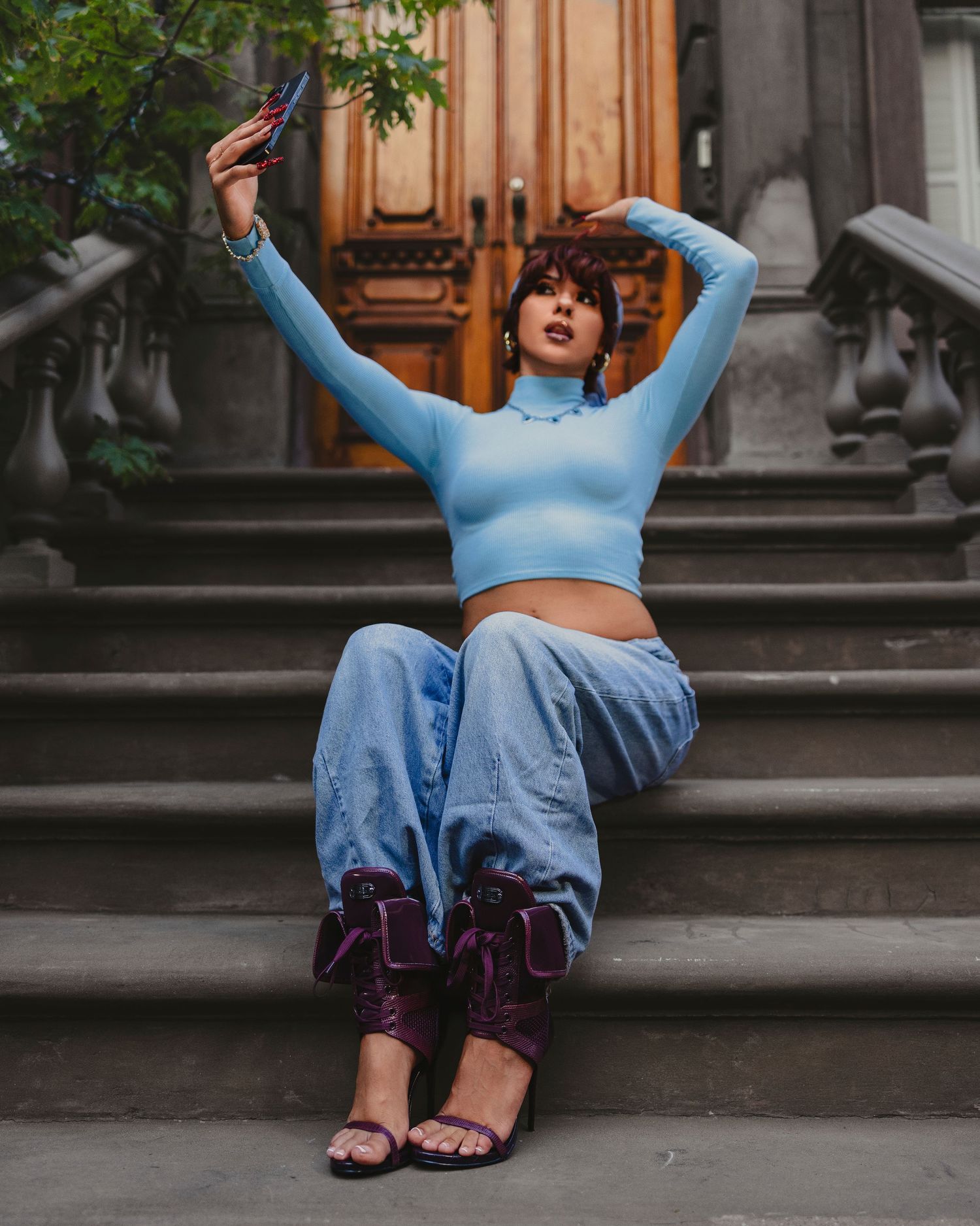 Woman in blue crop top and baggy jeans sits on stone steps taking a selfie wearing purple heeled sandals.