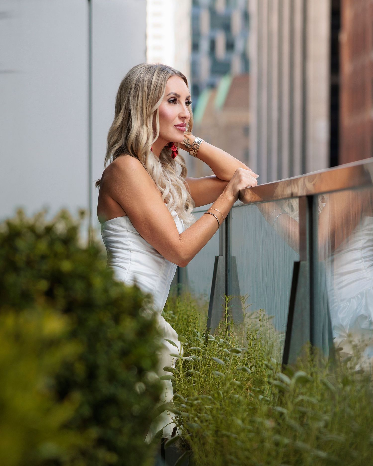 Blonde woman in white outfit leaning on a railing outdoors, surrounded by lush greenery in an urban setting.