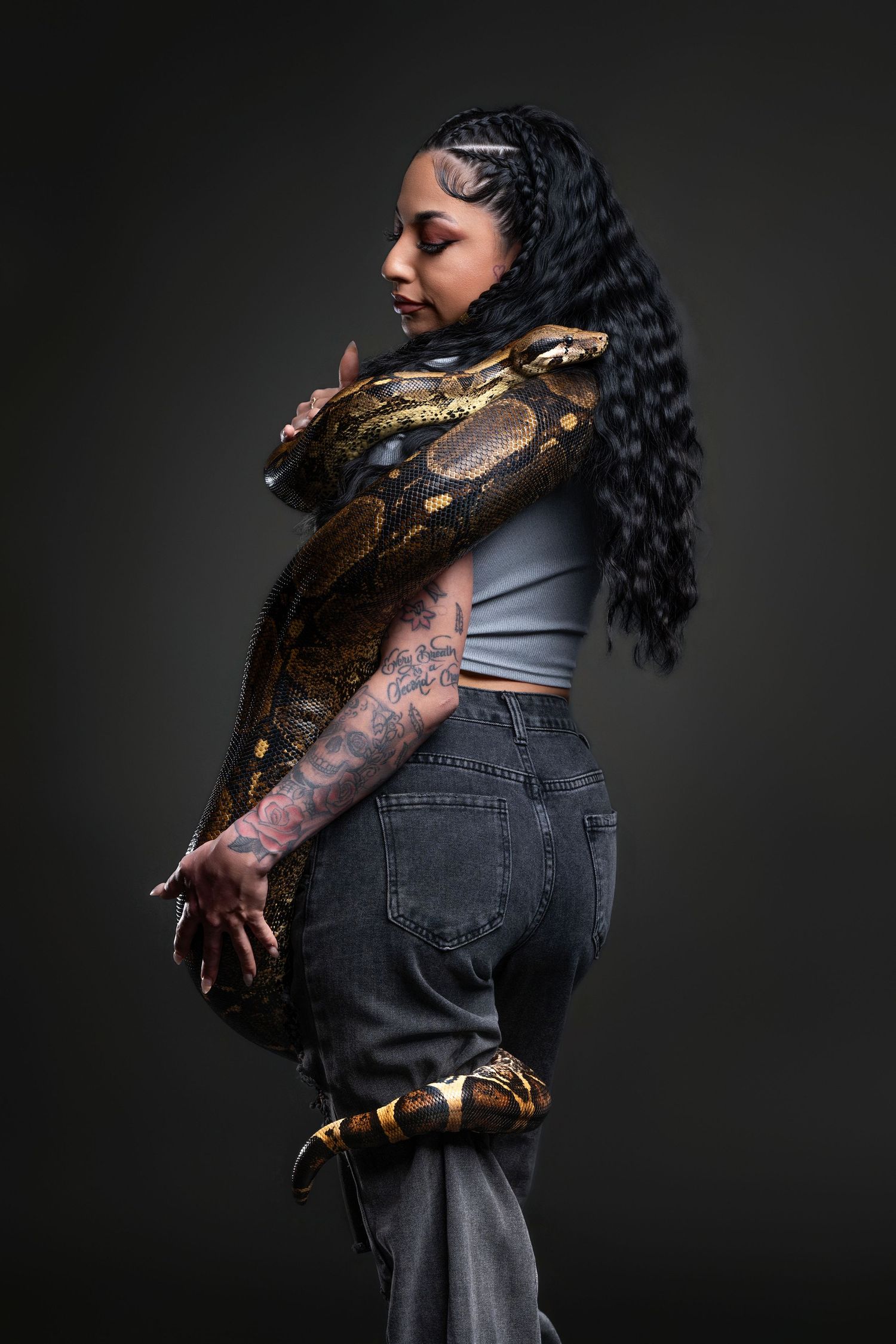 Woman with braids and tattoos poses with a large python snake draped over her shoulders in a studio.