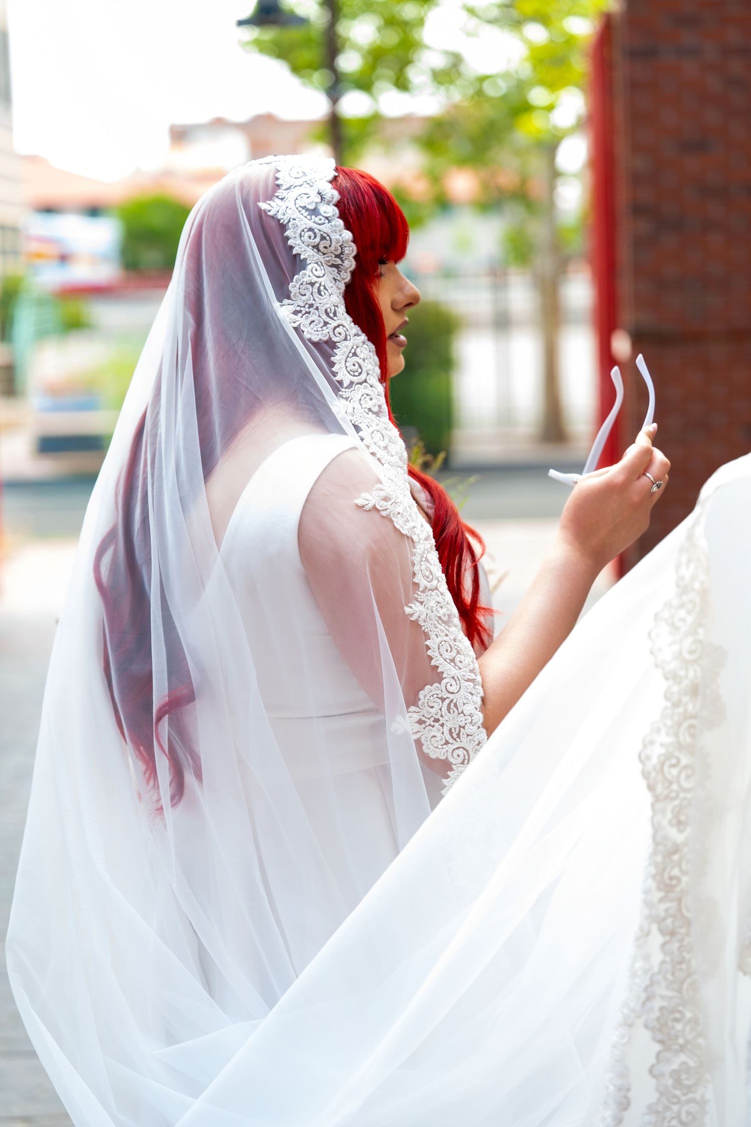 Red-haired bride in lace-trimmed veil checks phone outdoors on urban street before wedding ceremony.