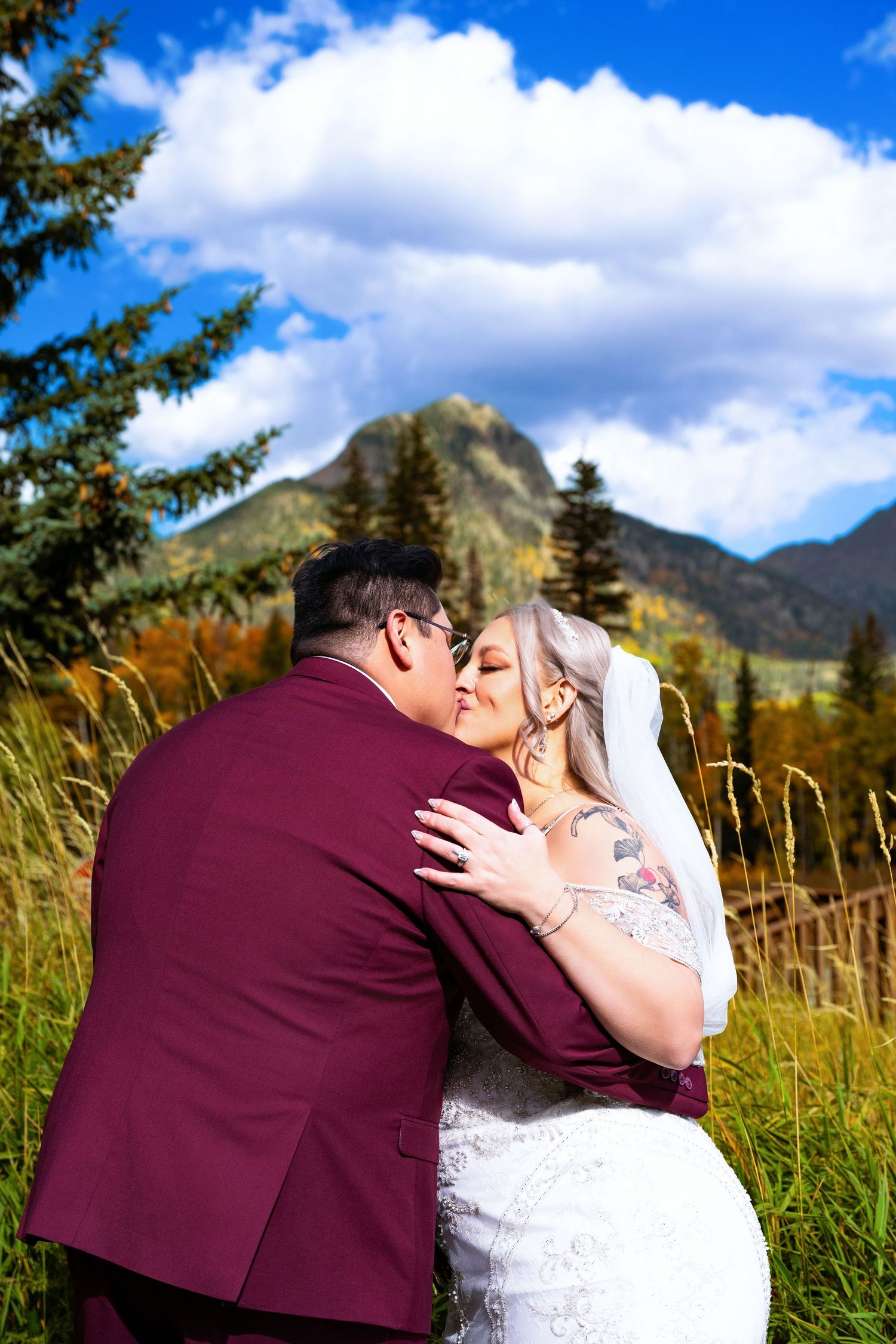 Couple shares romantic kiss in tall grass field with mountain peaks and blue sky behind them.