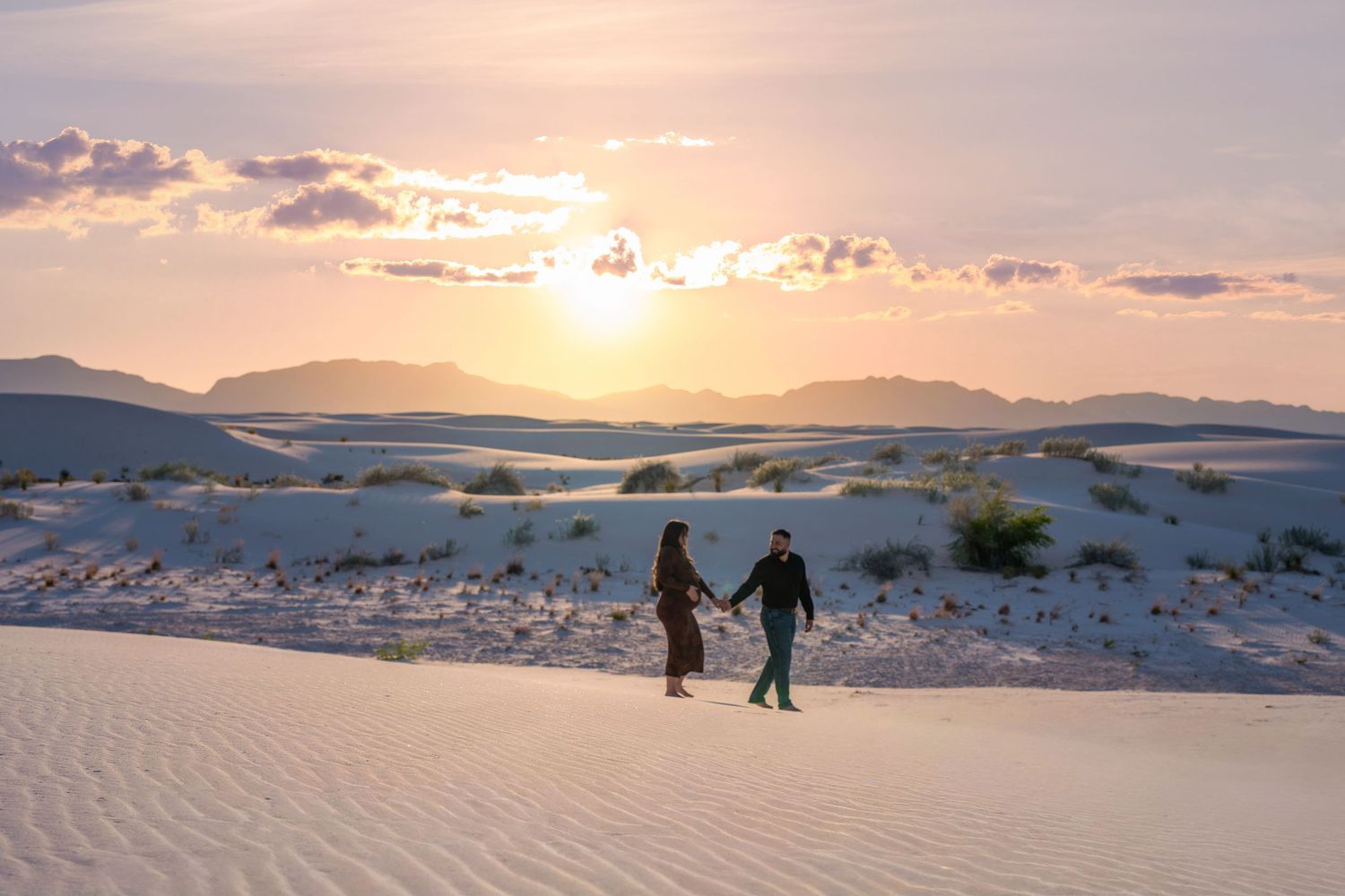 Couple holding hands walking across White Sands dunes at golden sunset with mountain backdrop.