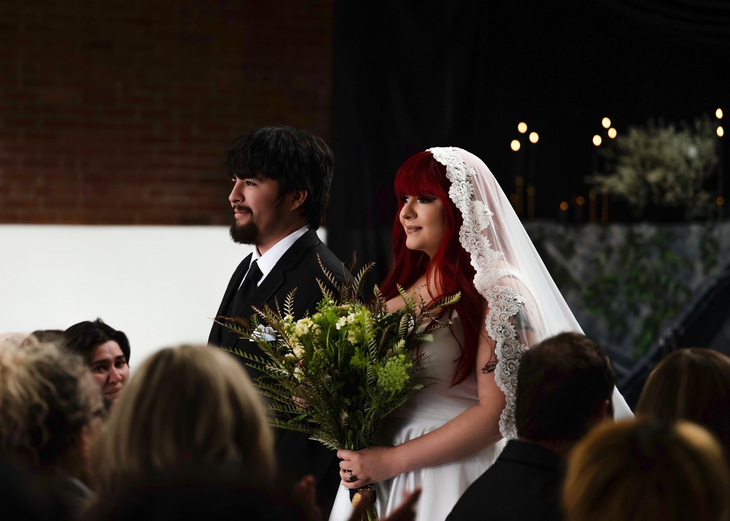 Red-haired bride in lace veil walks down aisle holding wildflower bouquet with groom at her side.
