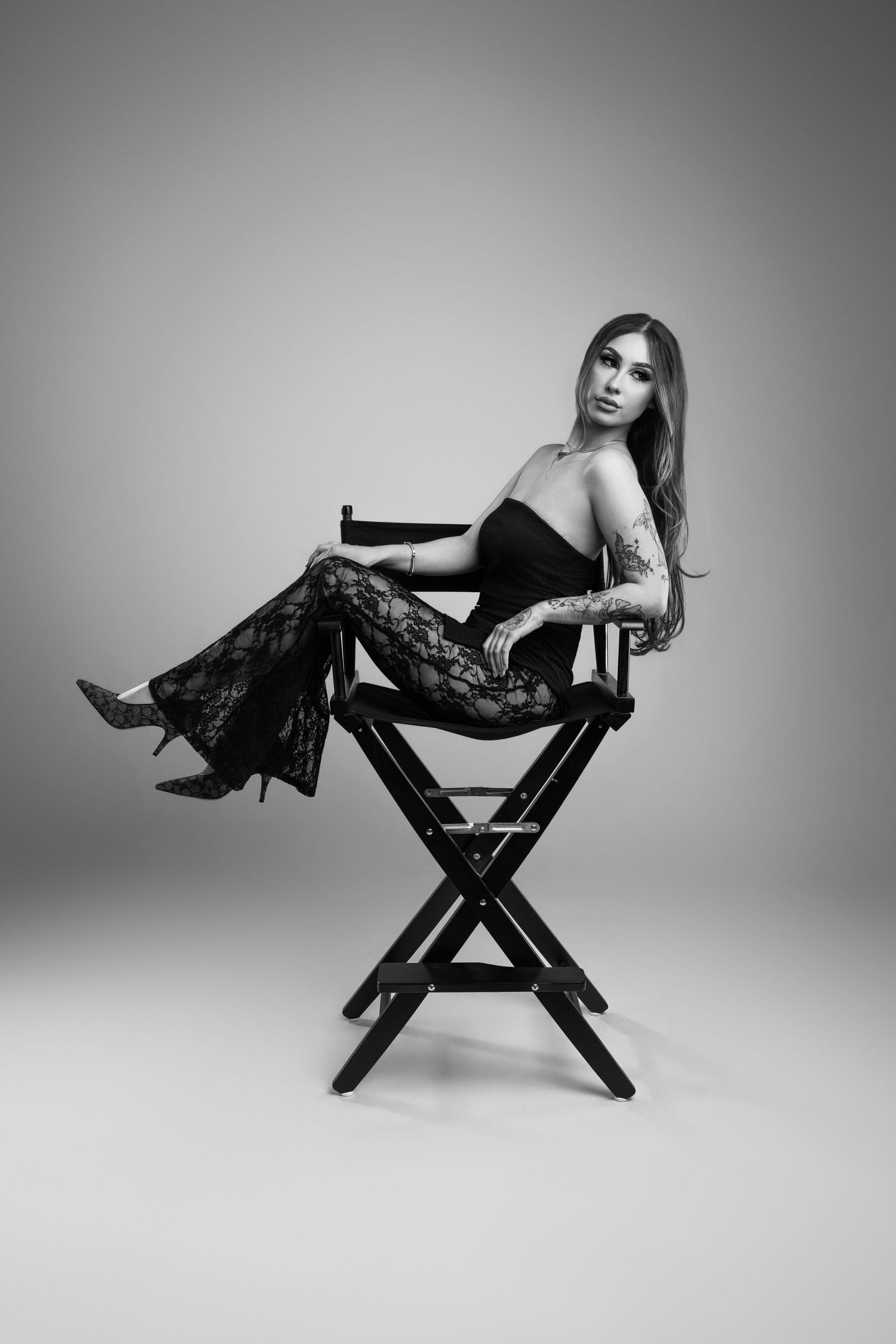 Black and white photo of tattooed woman in lace pants posing on director's chair in studio.