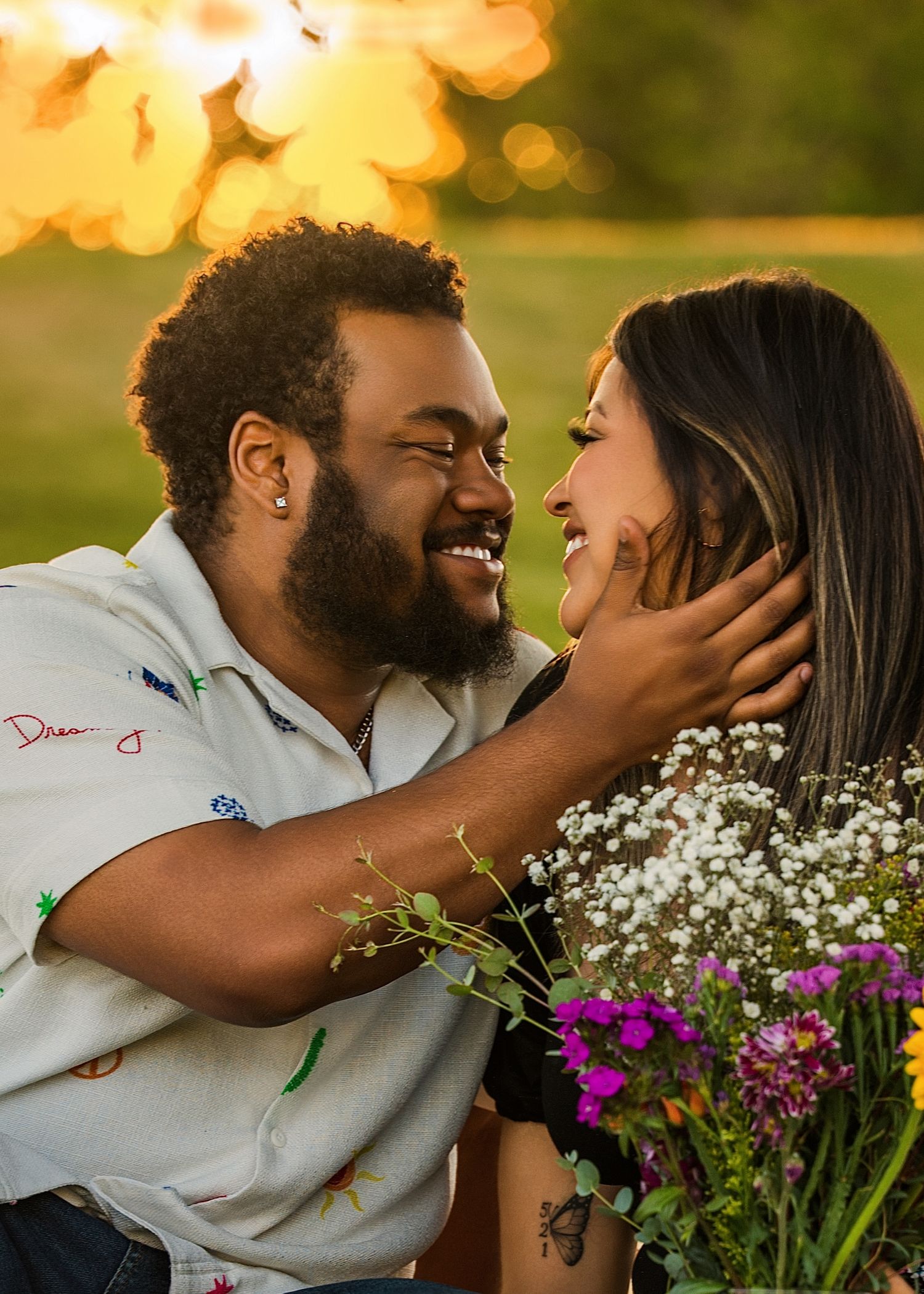 Smiling couple gazes at each other at golden sunset, man holds woman's face with wildflowers nearby.