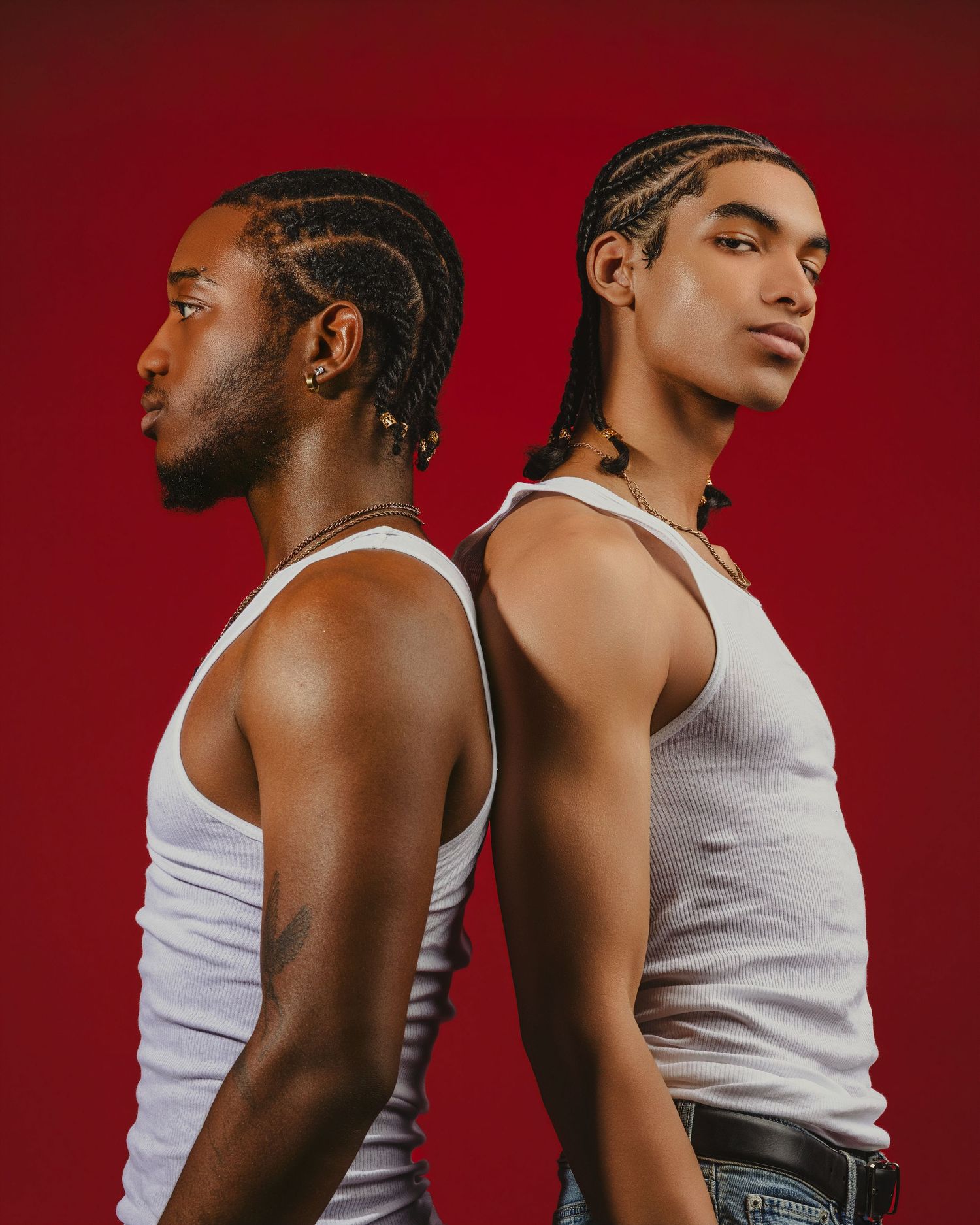 Two young men with braided hair wearing white tank tops pose back-to-back against a bold red background.