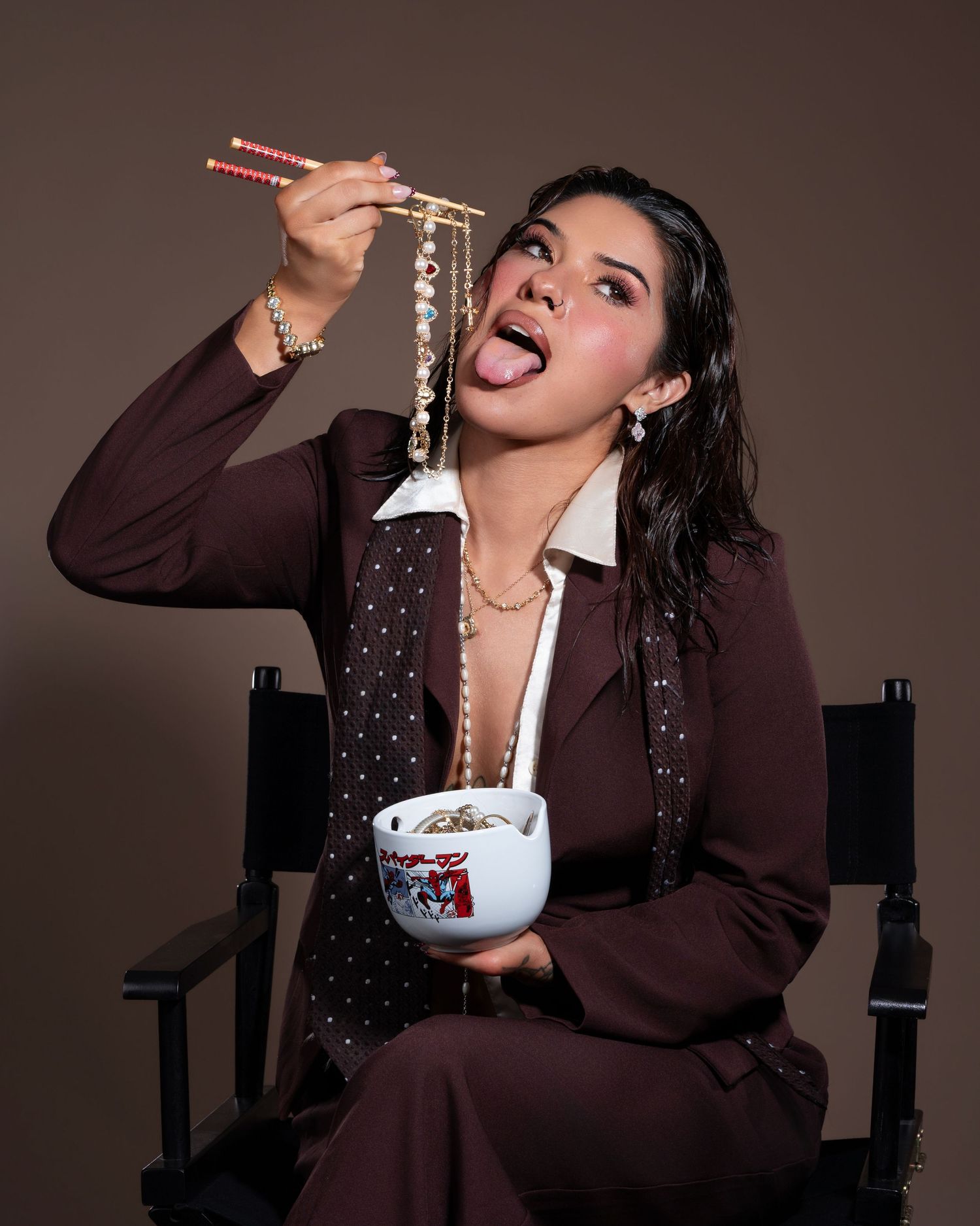 Woman in brown suit eating noodles with chopsticks from a bowl while sitting in a director's chair.