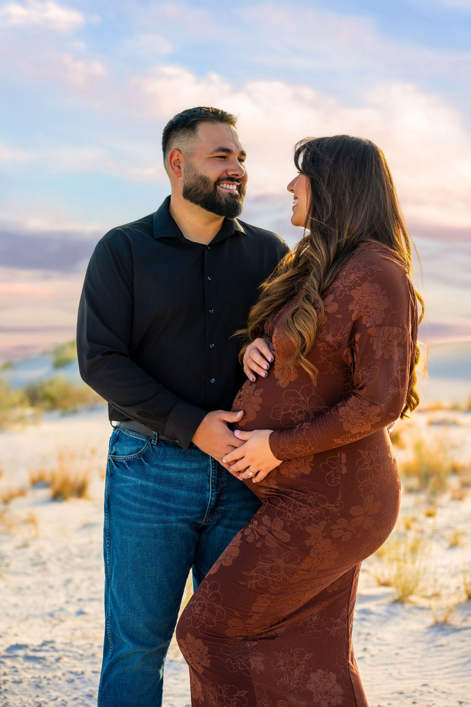 Couple sharing a joyful maternity moment on sand dunes at sunset, man in black shirt embracing pregnant woman in brown dress.