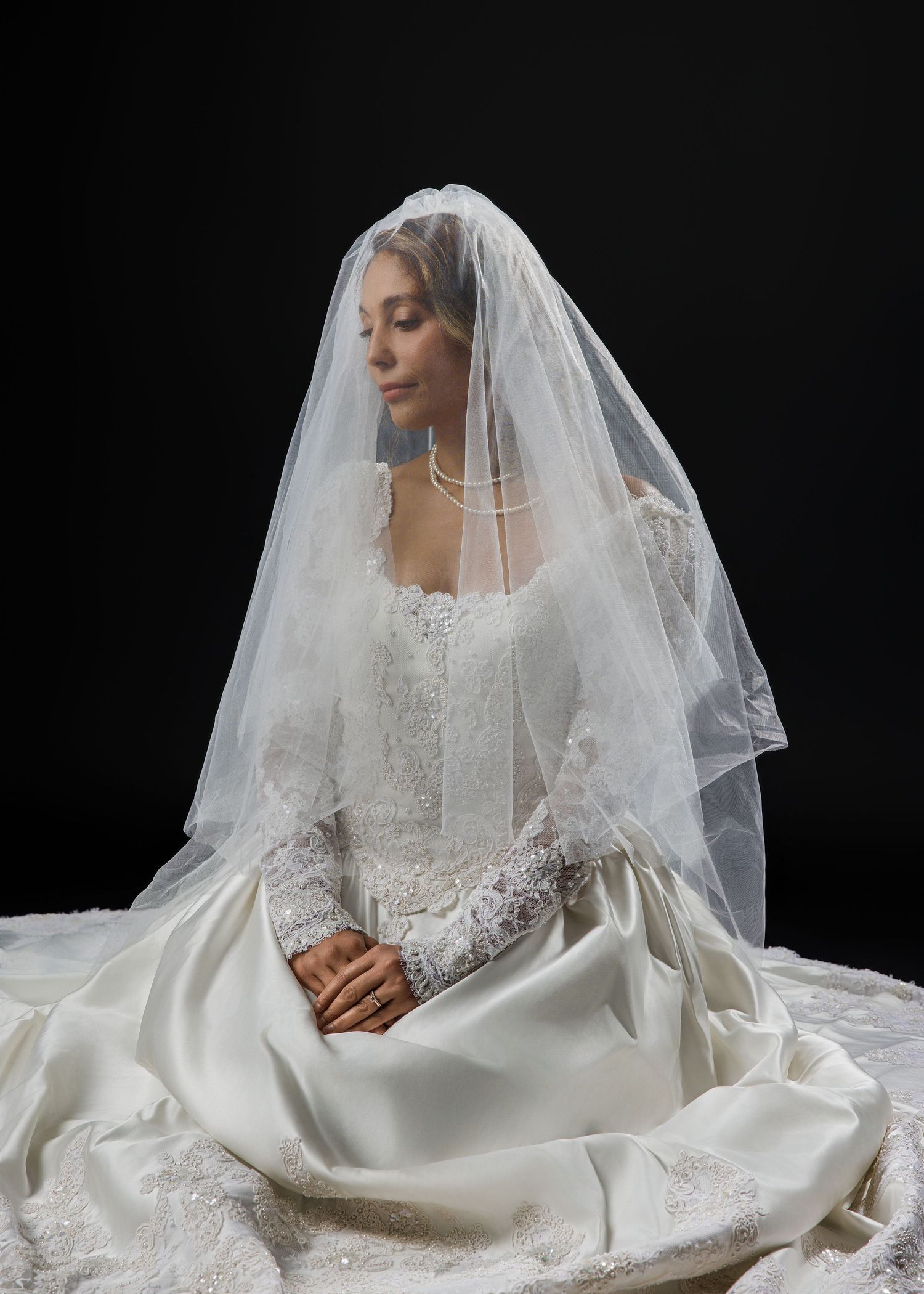 Bride in a lace wedding gown and veil sitting gracefully with eyes closed against a dark background.