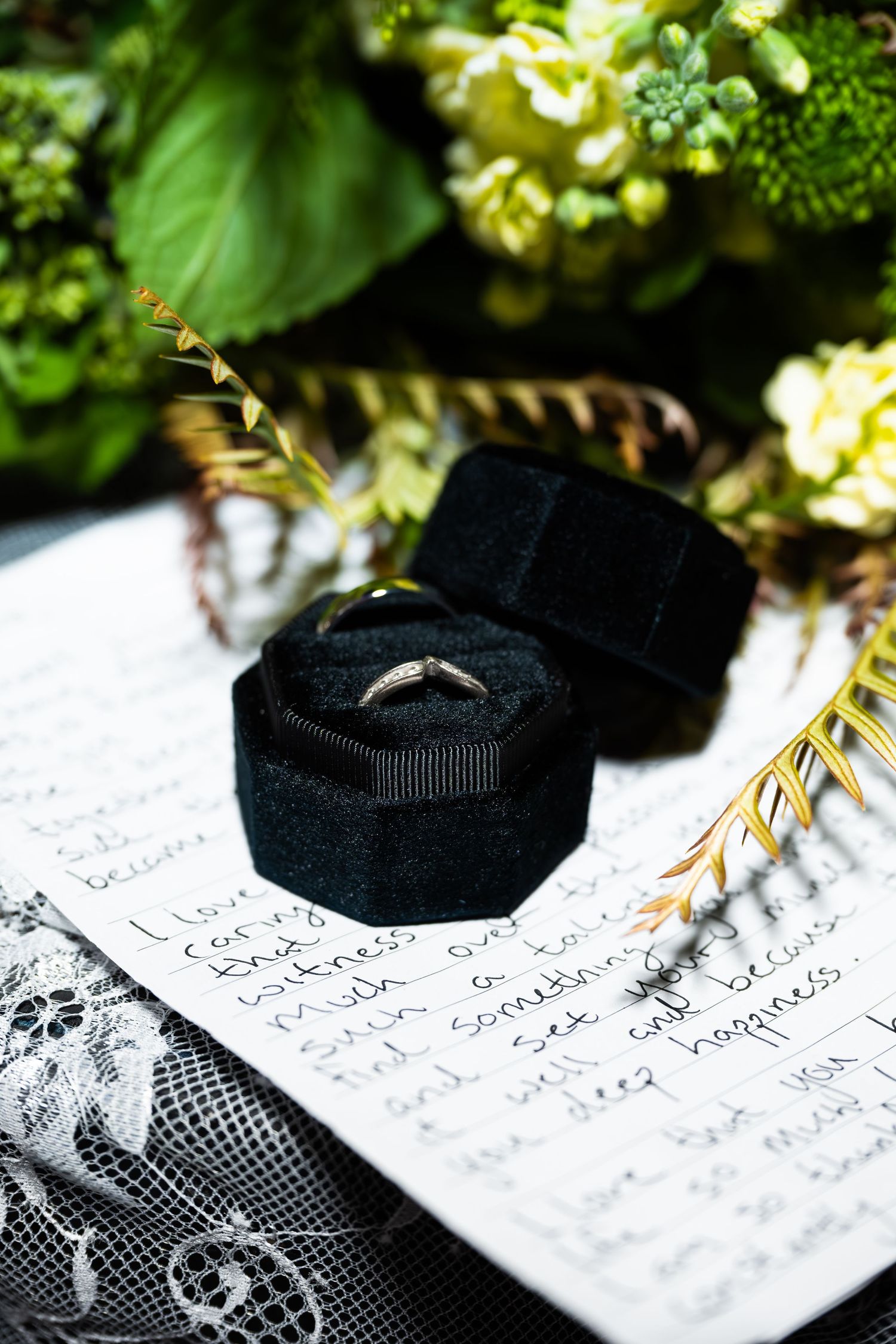 Diamond wedding ring in black velvet box resting on handwritten vows with green floral backdrop.