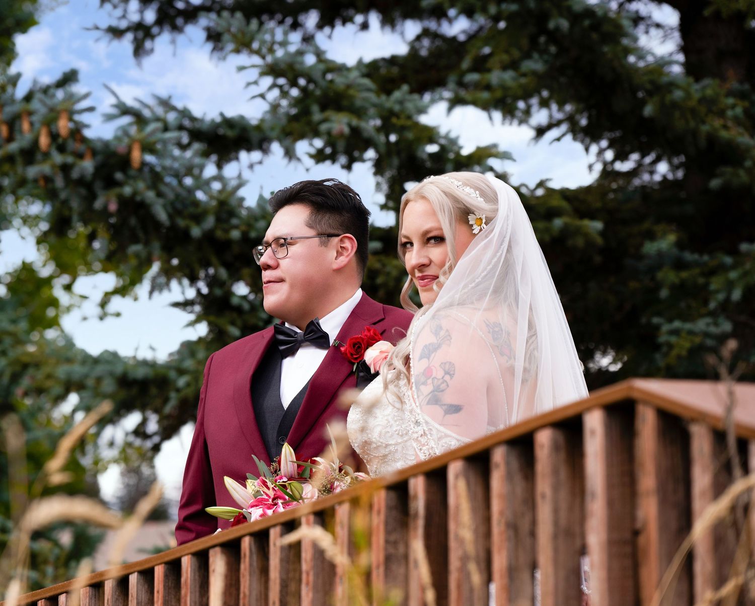 Bride and groom in burgundy suit stand on wooden deck surrounded by evergreen trees.