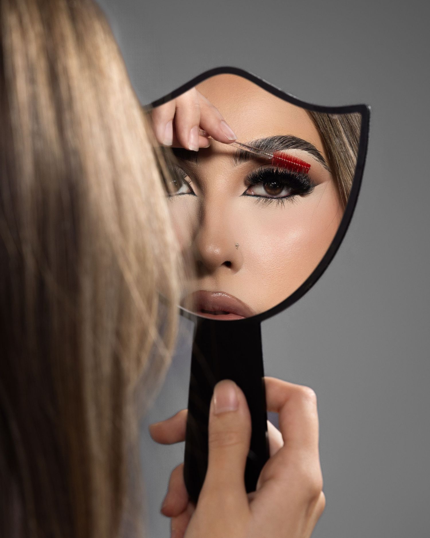 Woman applying makeup while holding a black hand mirror, showcasing bold red and black eye makeup with defined brows.