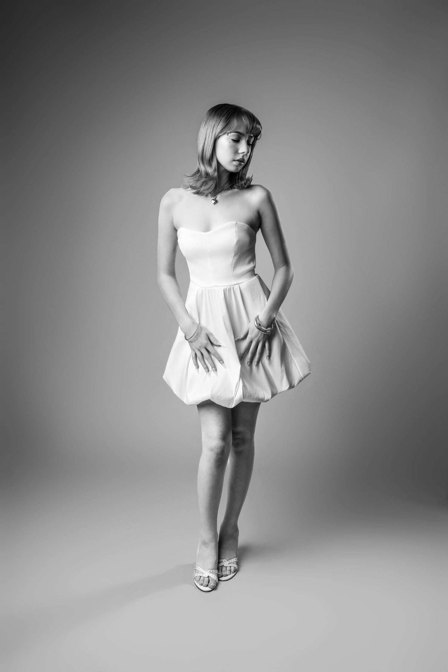 Black and white studio photo of a young woman in a strapless bubble mini dress and heels posing gracefully.