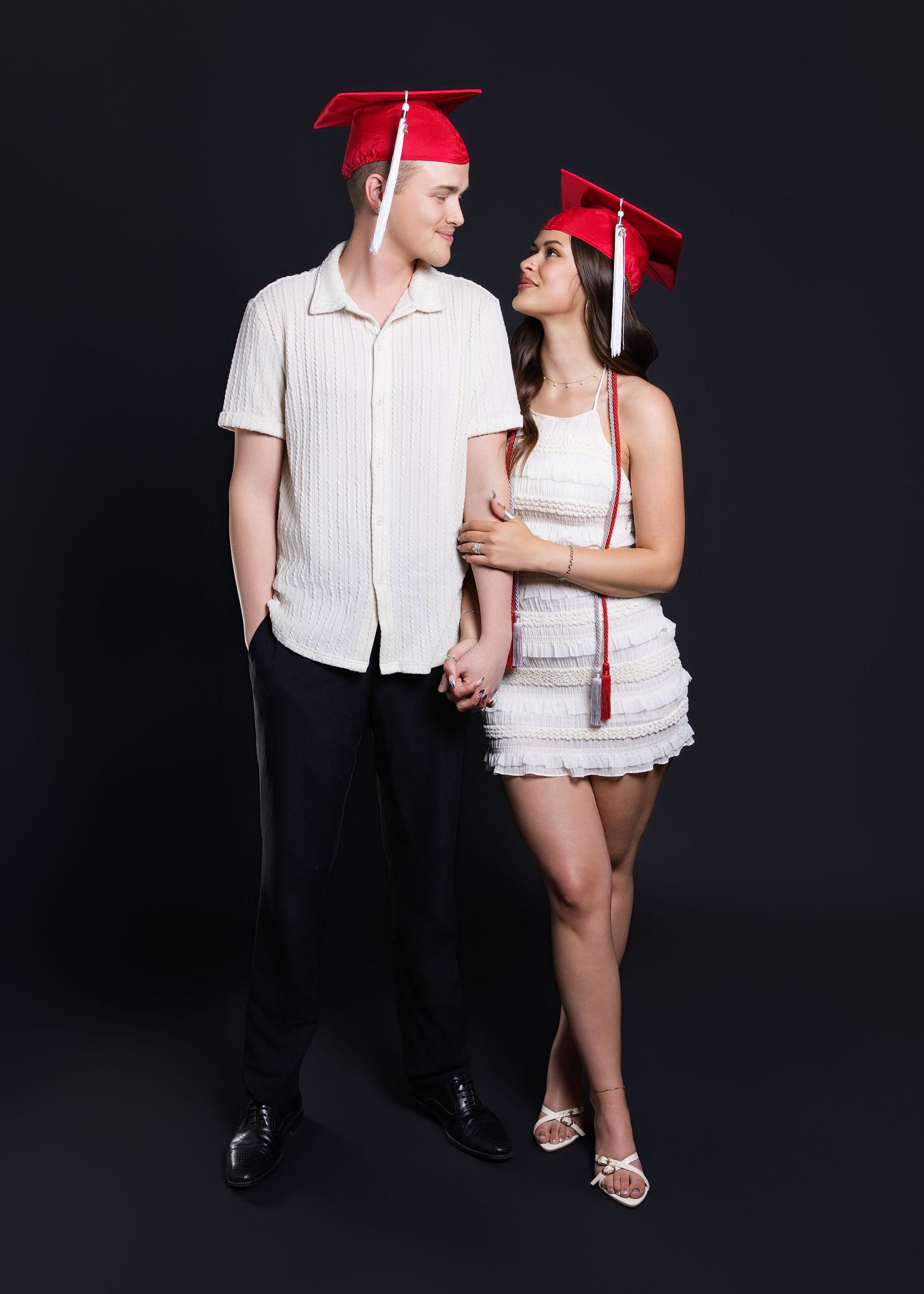 Young couple in red graduation caps posing together in a dark studio, looking at each other.