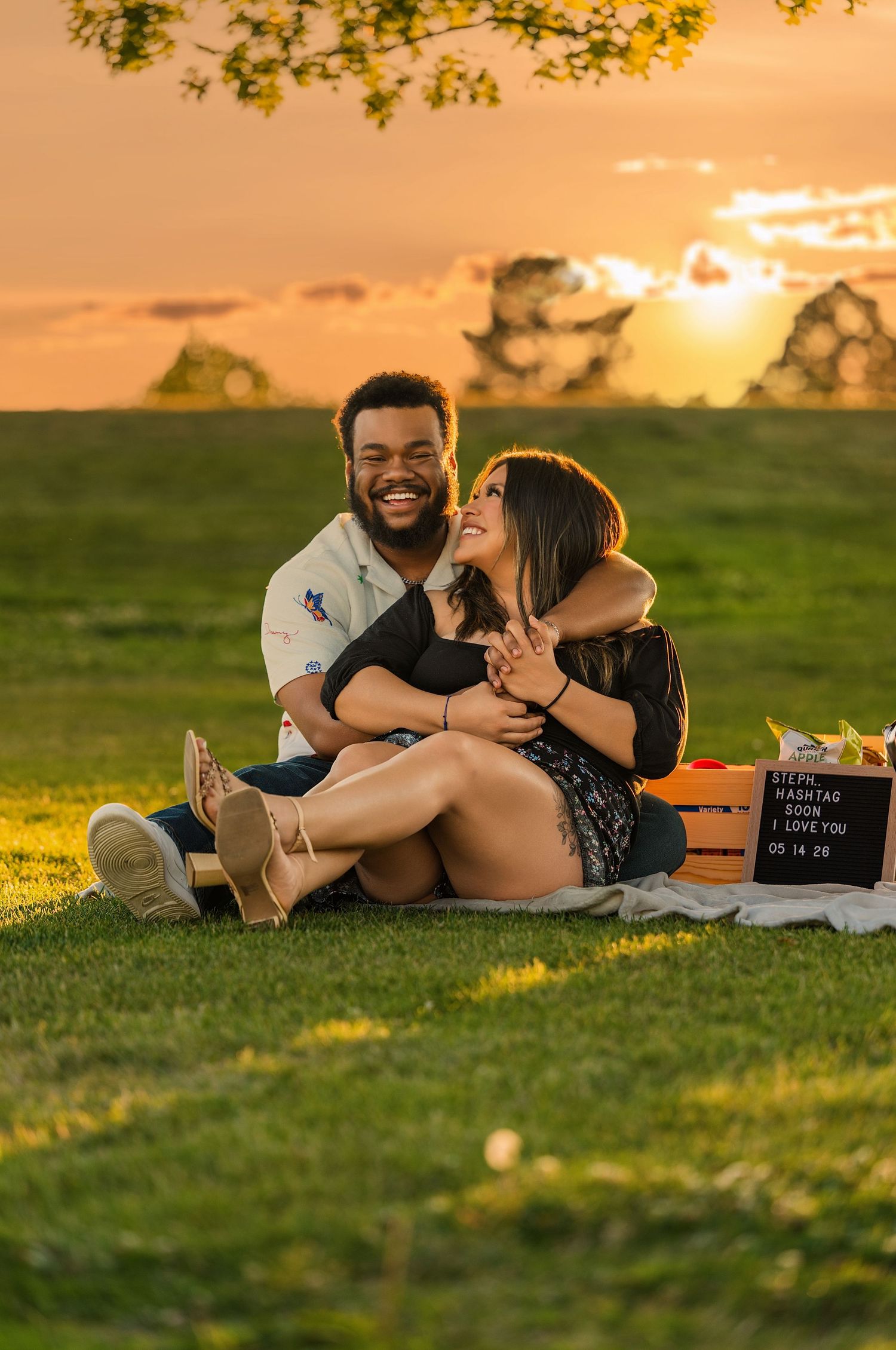 Happy couple sits together on grass at golden sunset during romantic outdoor engagement photo session.