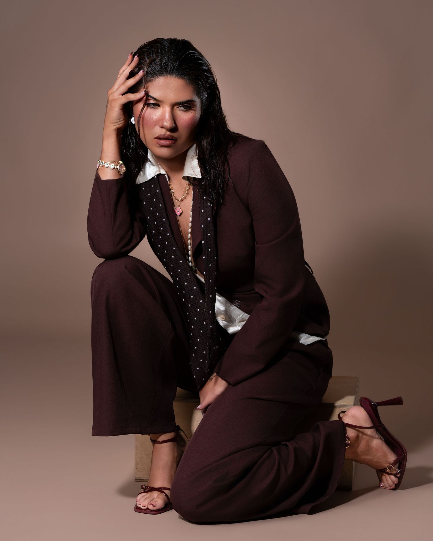 Woman in brown suit and heeled sandals poses seated on wooden block against neutral background.