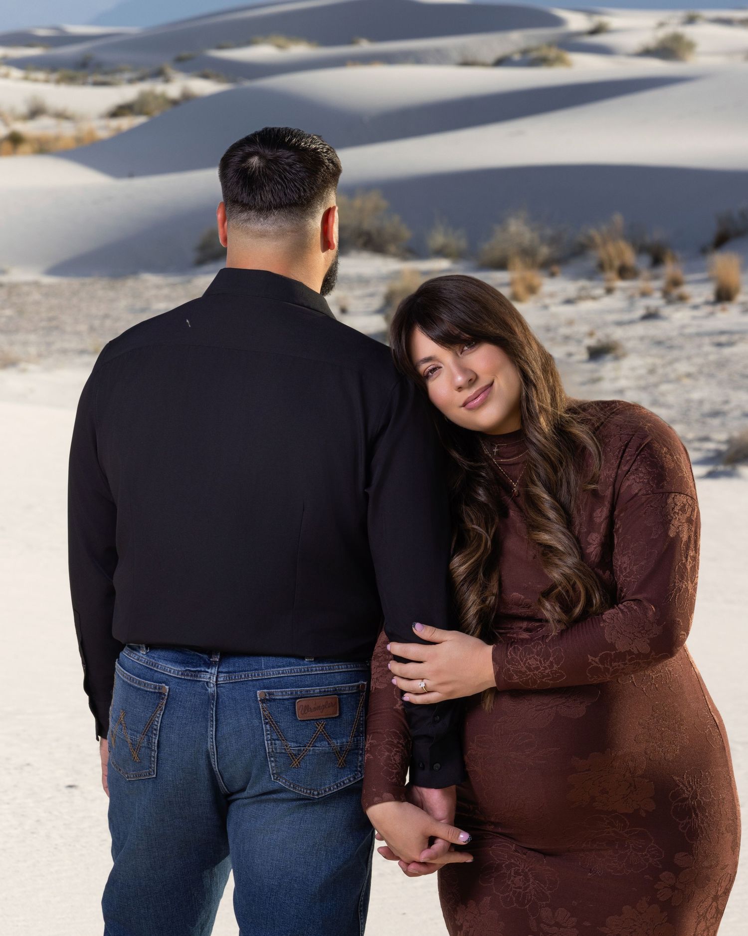 Pregnant woman in brown lace dress leaning on partner at White Sands National Park at dusk.