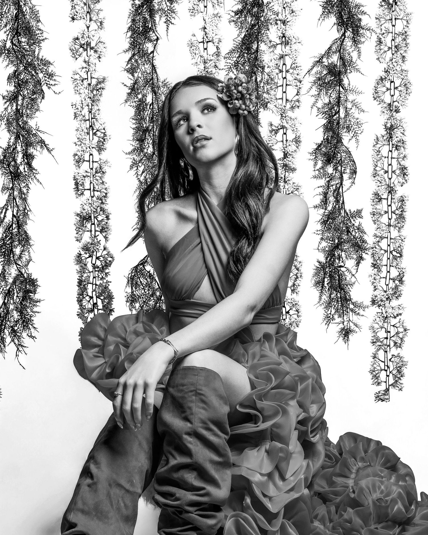 Black and white photo of a woman in a ruffled gown with floral crown, posed against hanging greenery backdrop.