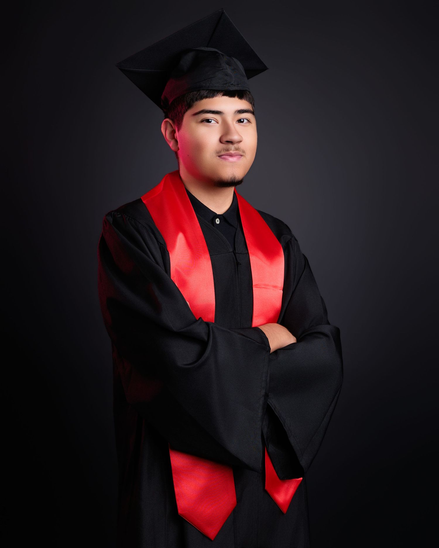Graduate posing with arms crossed wearing black cap, gown, and red sash in studio portrait.