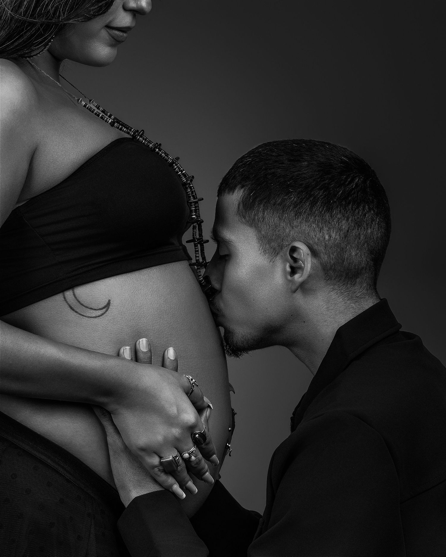 Black and white photo of partner kissing pregnant belly with rosary beads in studio setting.