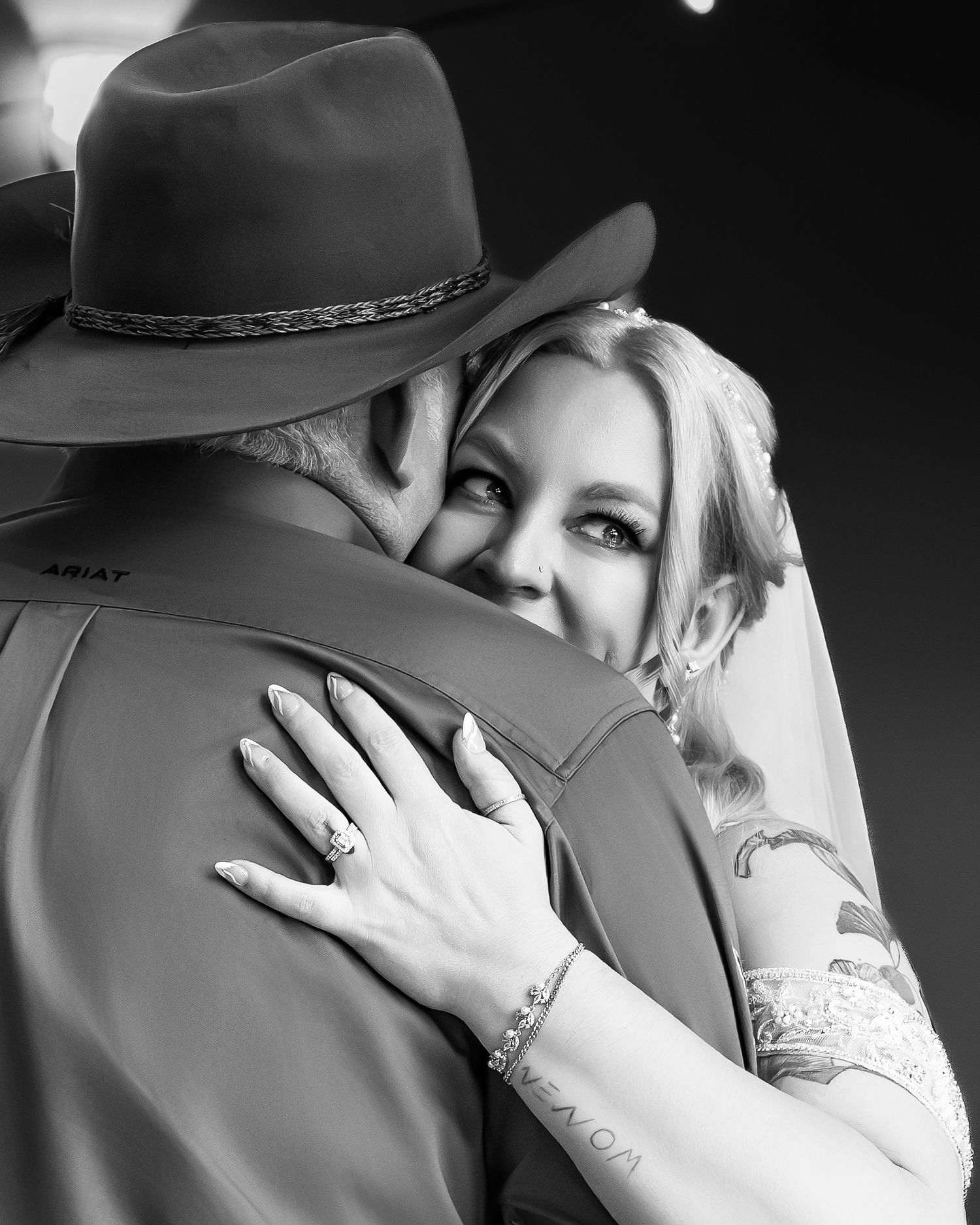 Bride with tattoos embracing groom in cowboy hat, showing wedding ring in black and white photo.