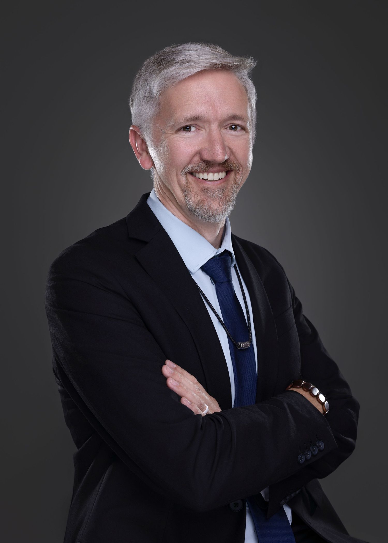 Smiling silver-haired businessman in dark suit and blue tie posing with arms crossed on gray background.