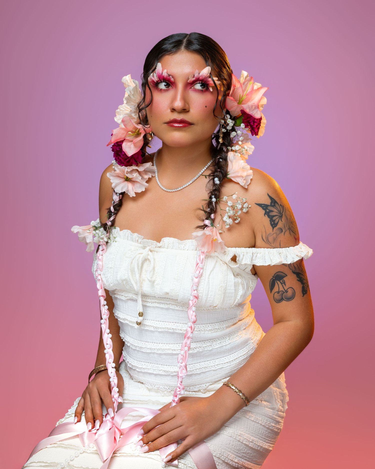 Woman in white corset dress with floral hair accessories, pink eye makeup, and tattoos poses against pink background.