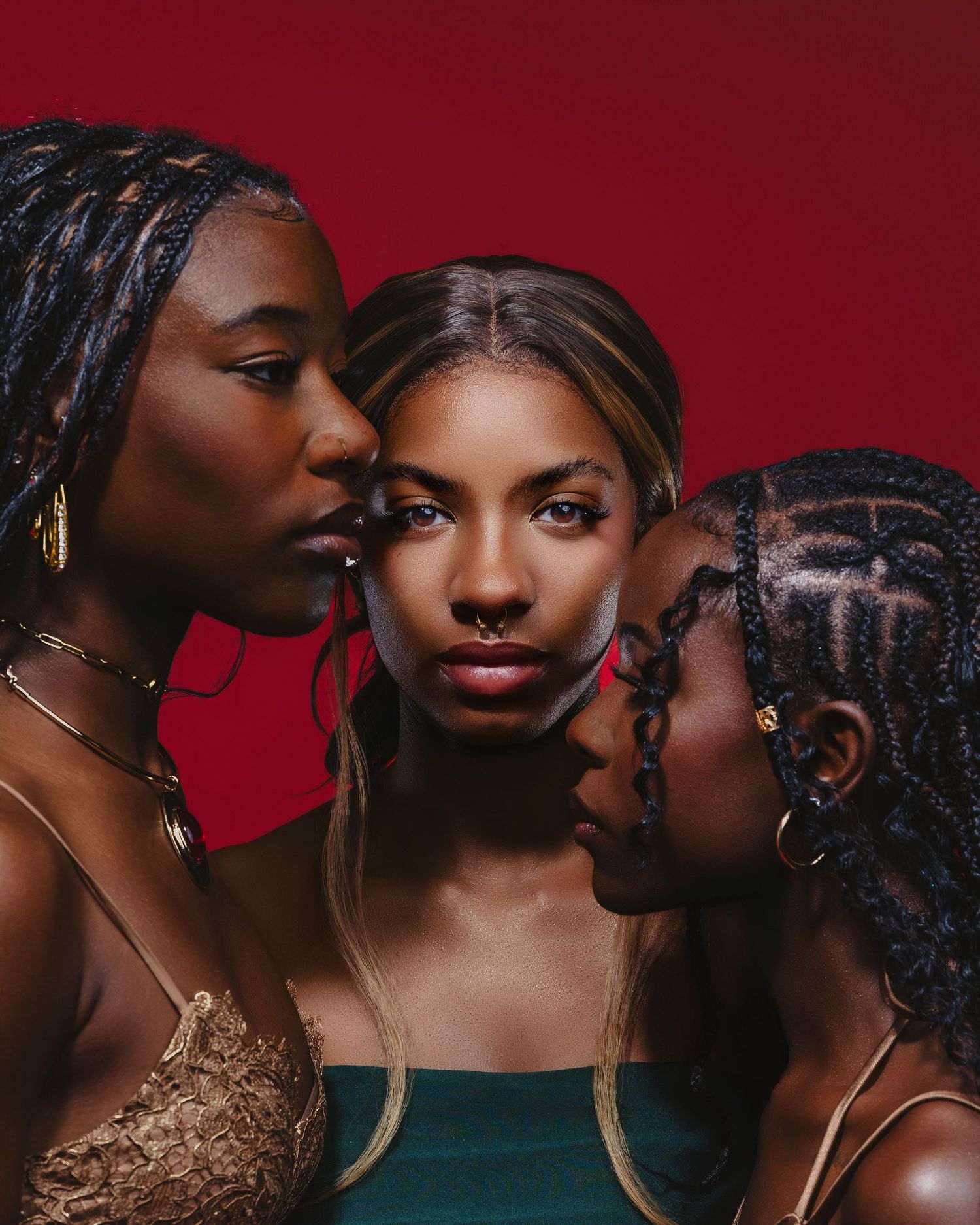 Three young Black women with braids and gold jewelry pose closely together against a bold red background.