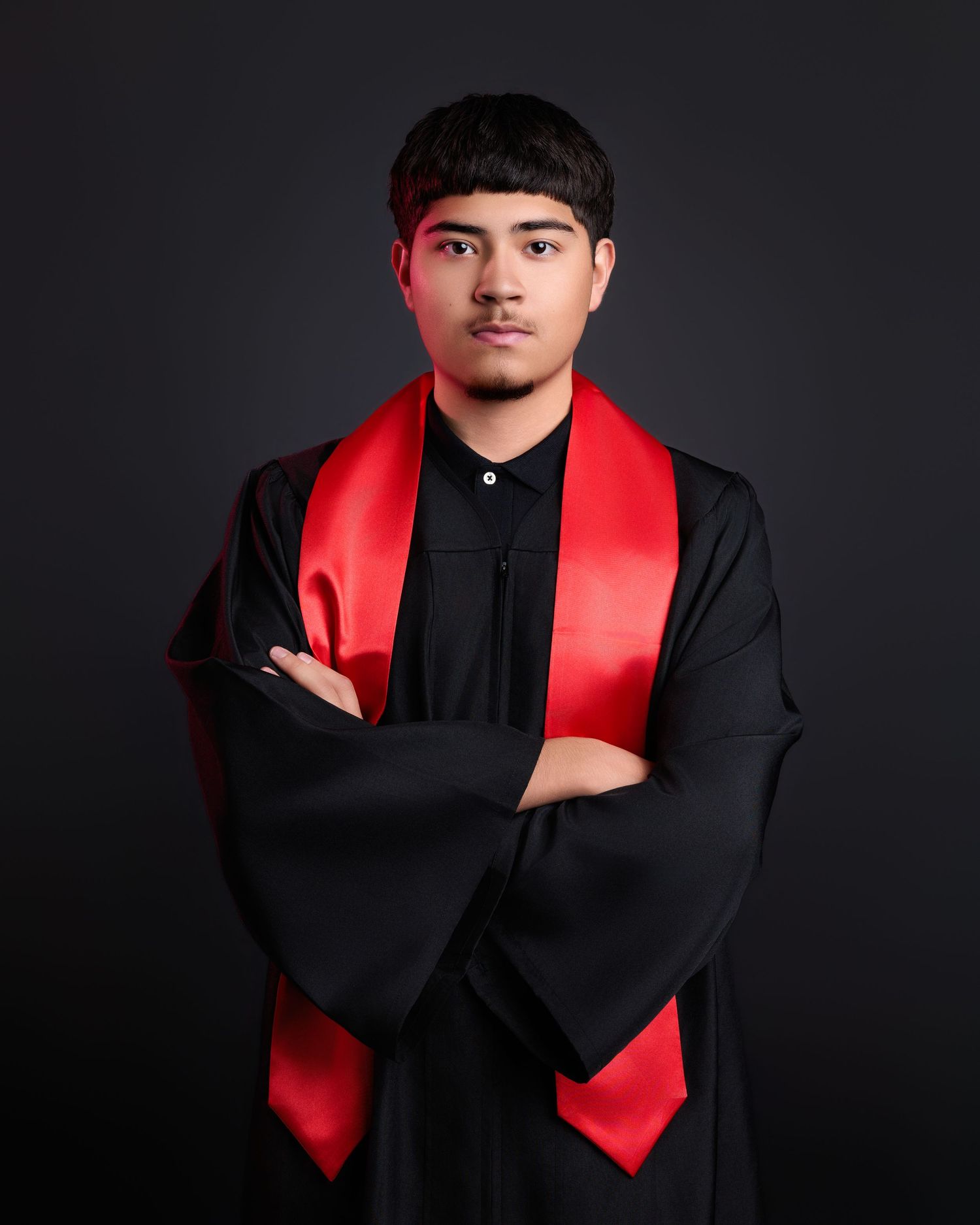 Graduate posing with arms crossed wearing black gown and red sash against dark background.