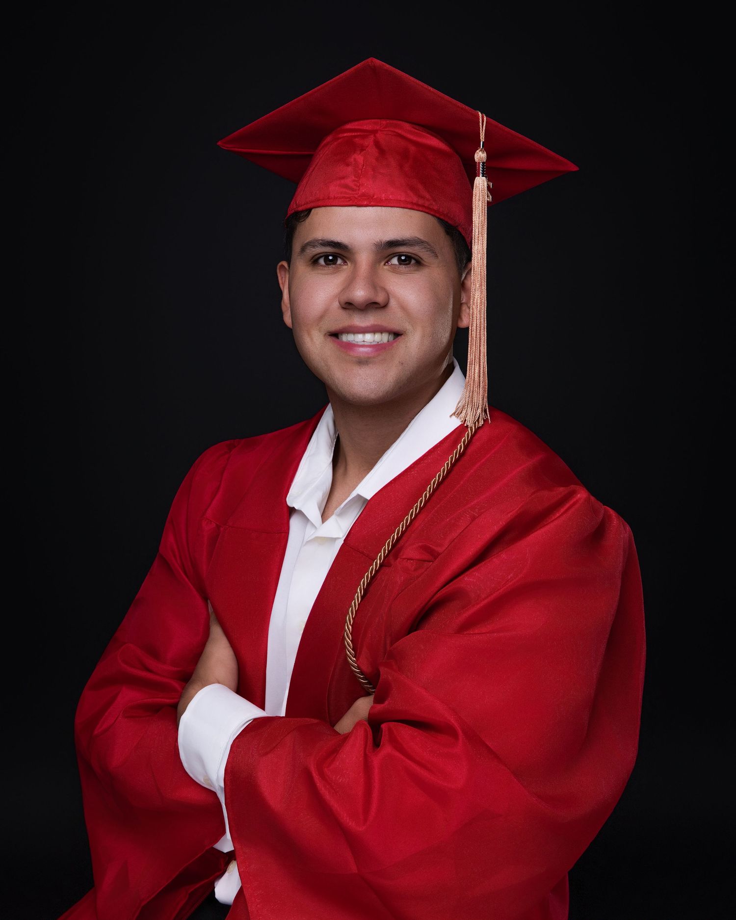 Smiling male graduate in red cap and gown with gold tassel posing with arms crossed in studio.