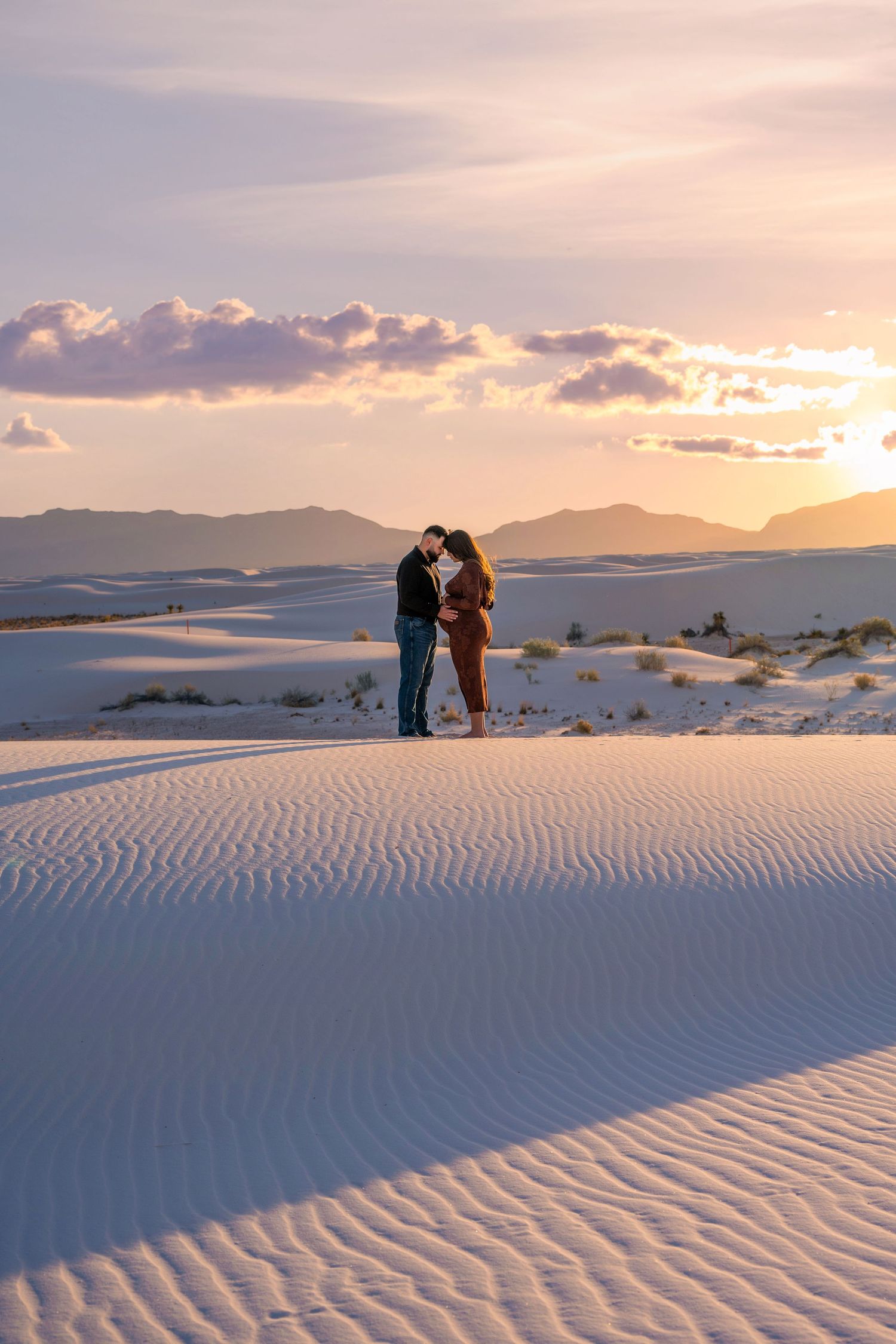 Couple sharing a kiss on White Sands dunes at sunset, woman in brown maternity dress.
