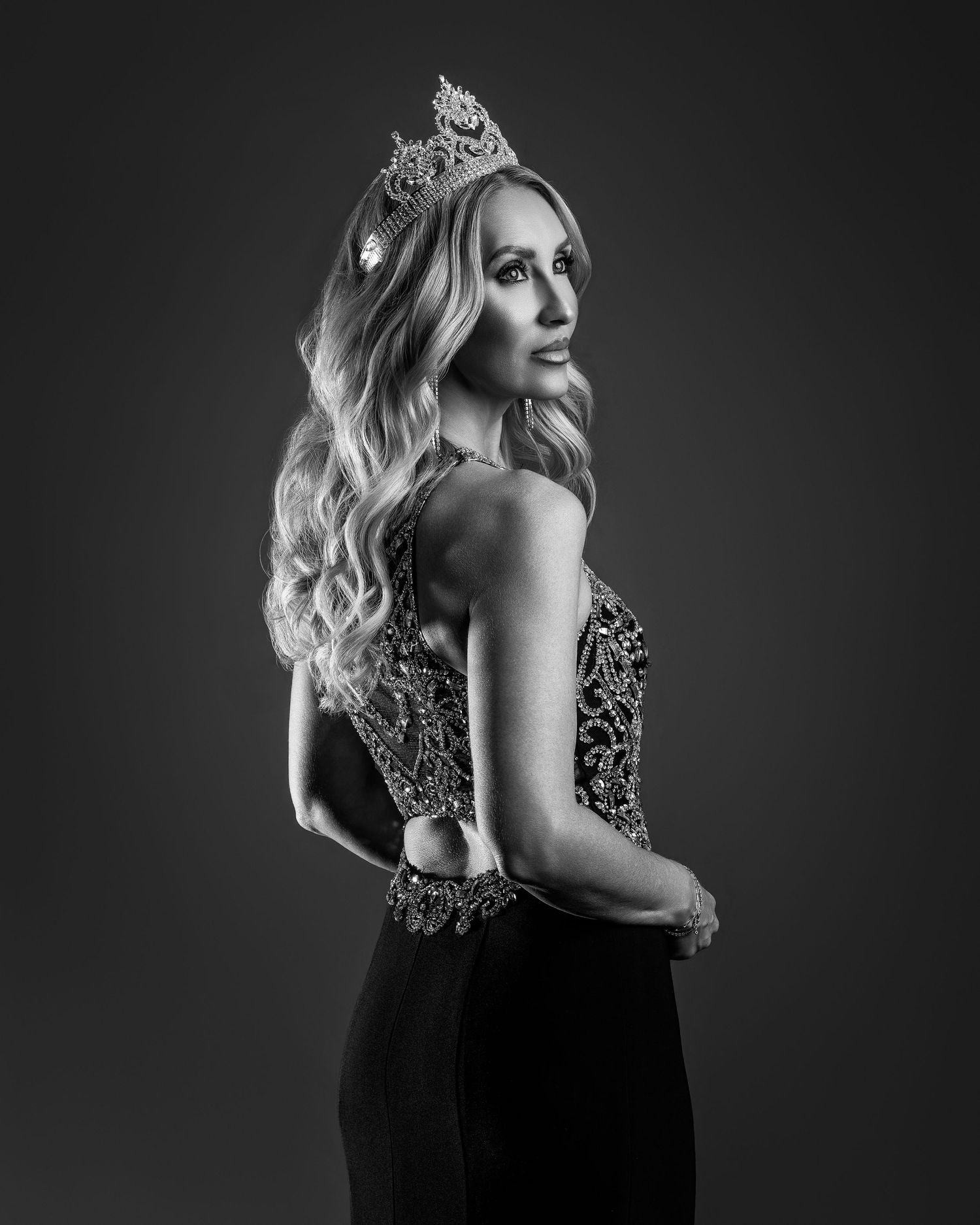 Black and white portrait of a woman in an elegant gown and ornate crown, posing gracefully in a studio.