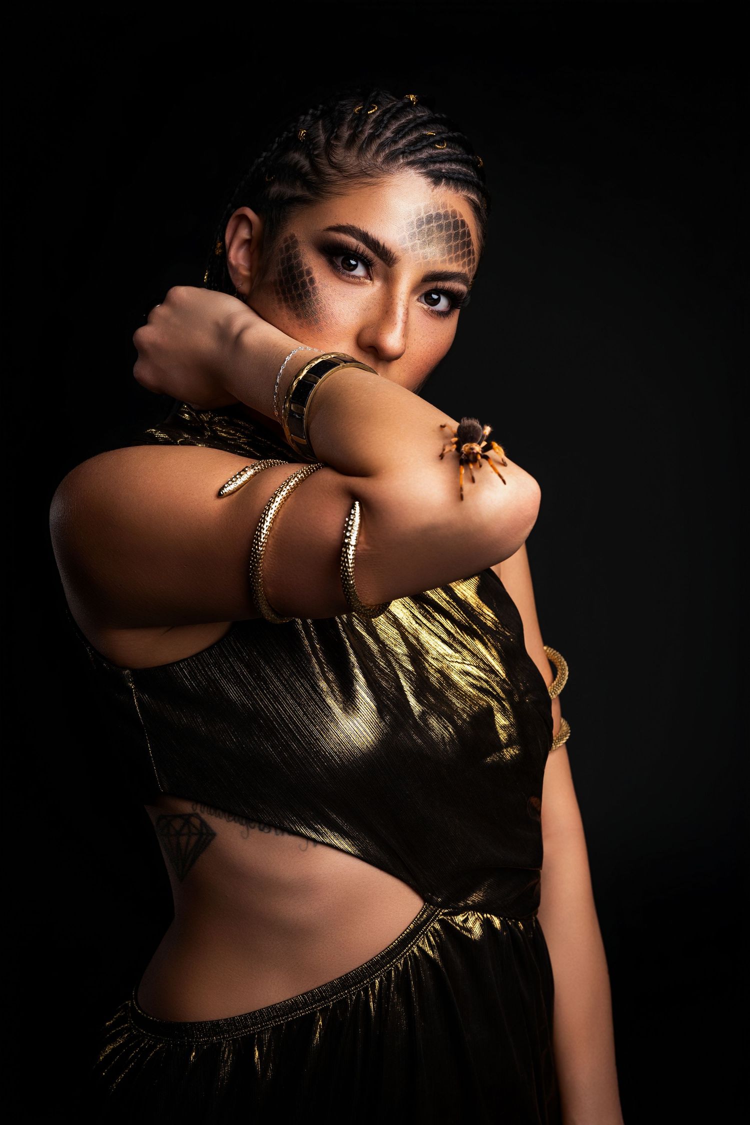 Woman in gold costume with braided hair and tarantula on arm poses dramatically against black background.