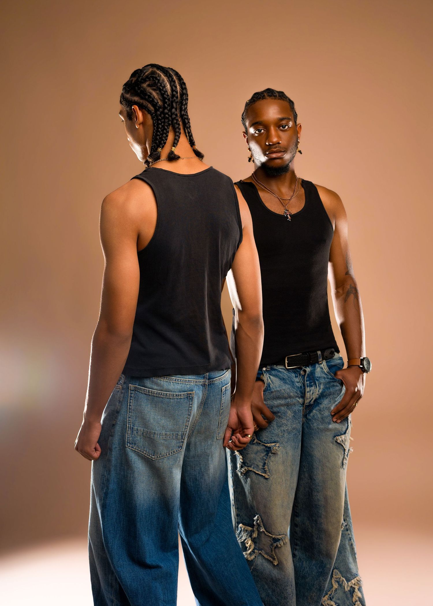 Two young men in black tank tops and baggy distressed jeans pose back-to-back on a beige background.